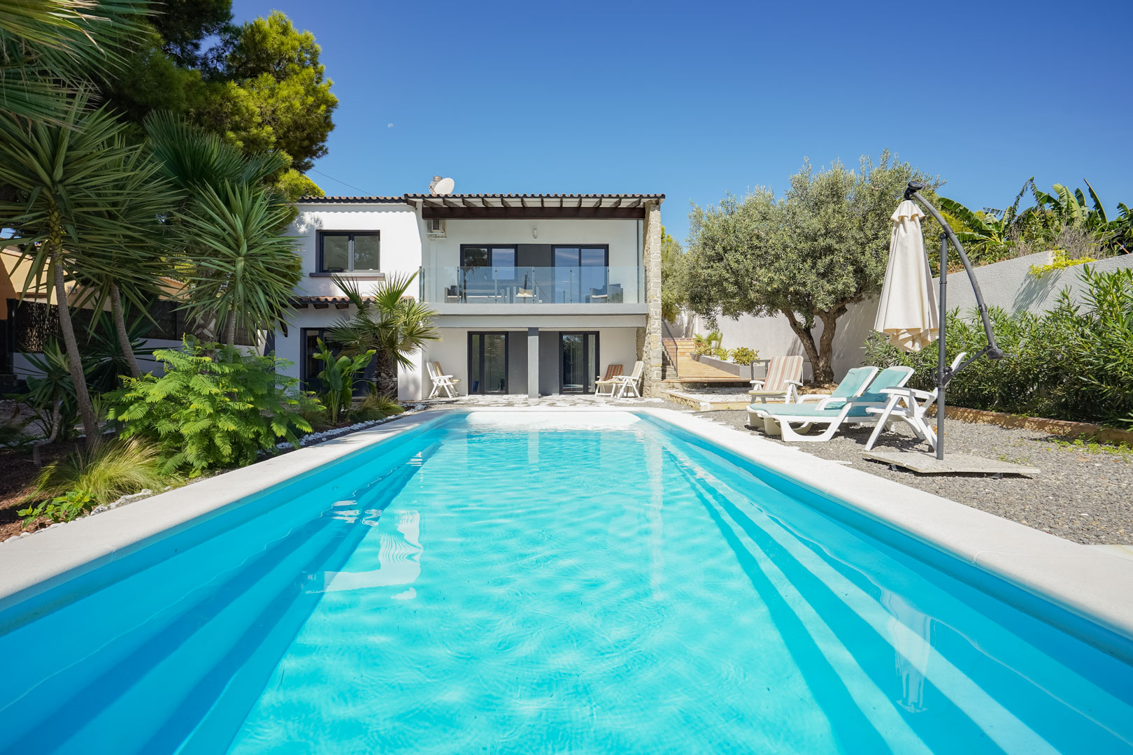 Blick auf den rechteckigen Pool und den mediterranen Garten mit Palmen, die weiße Fassade der Villa und die obere Terrasse mit Glasgeländer zeigend.