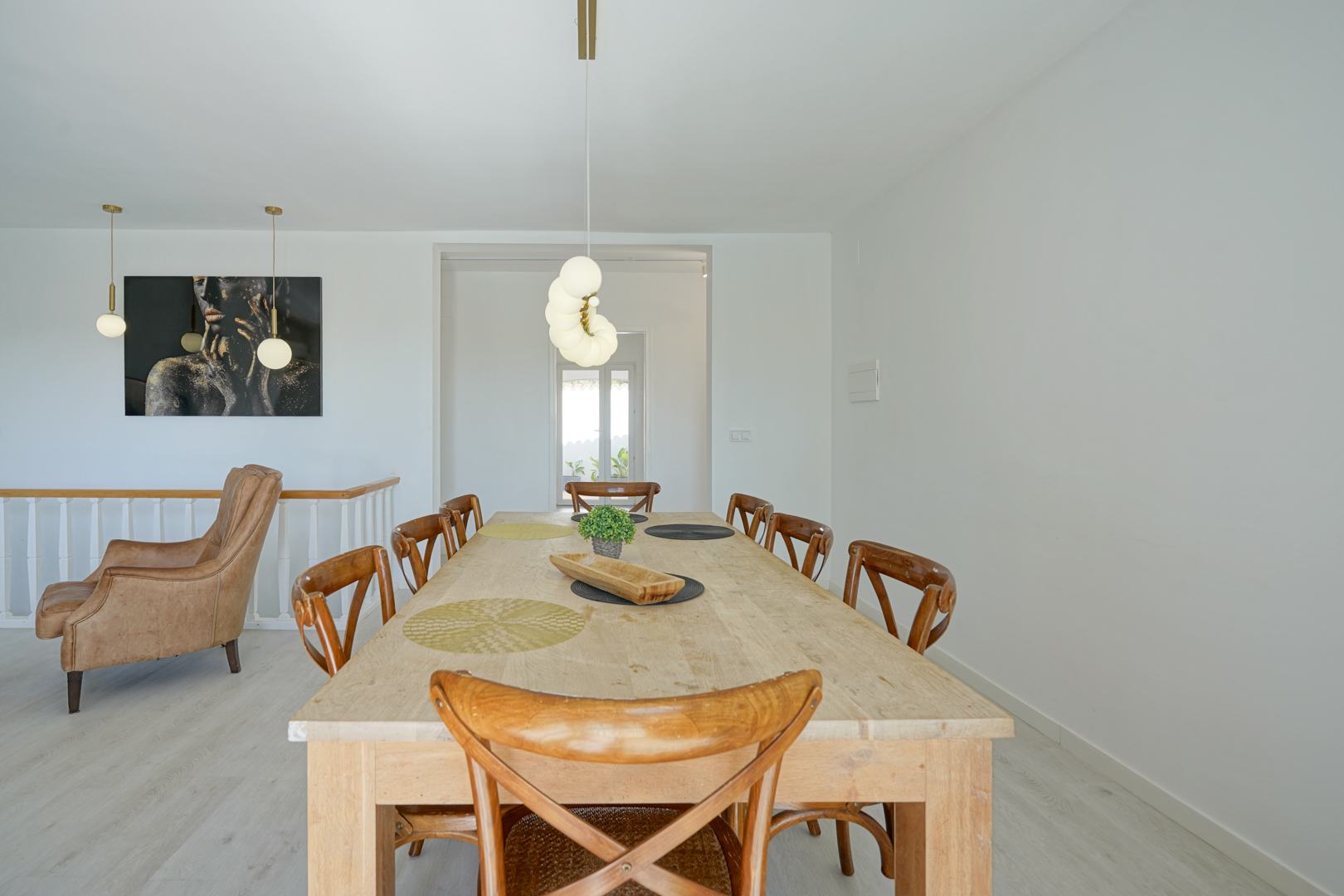 Essbereich mit einem großen hellen Holztisch, X-Back-Stühlen und modernen Kugelpendelleuchten. Im Hintergrund befinden sich ein Ledersessel und ein markantes Wandgemälde.