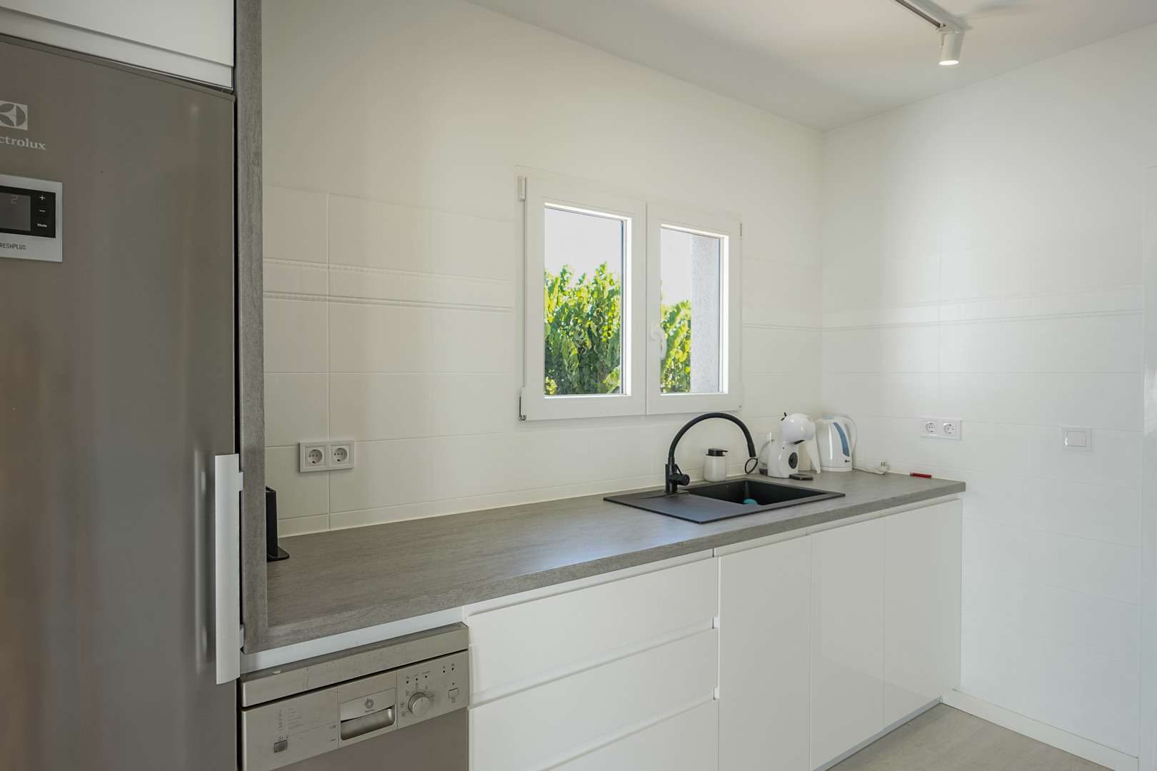 Funktionale Küche mit grifflosen weißen Schränken, grauer Arbeitsplatte, schwarzem Spülbecken und integrierten Geräten an einem hellen Fenster.