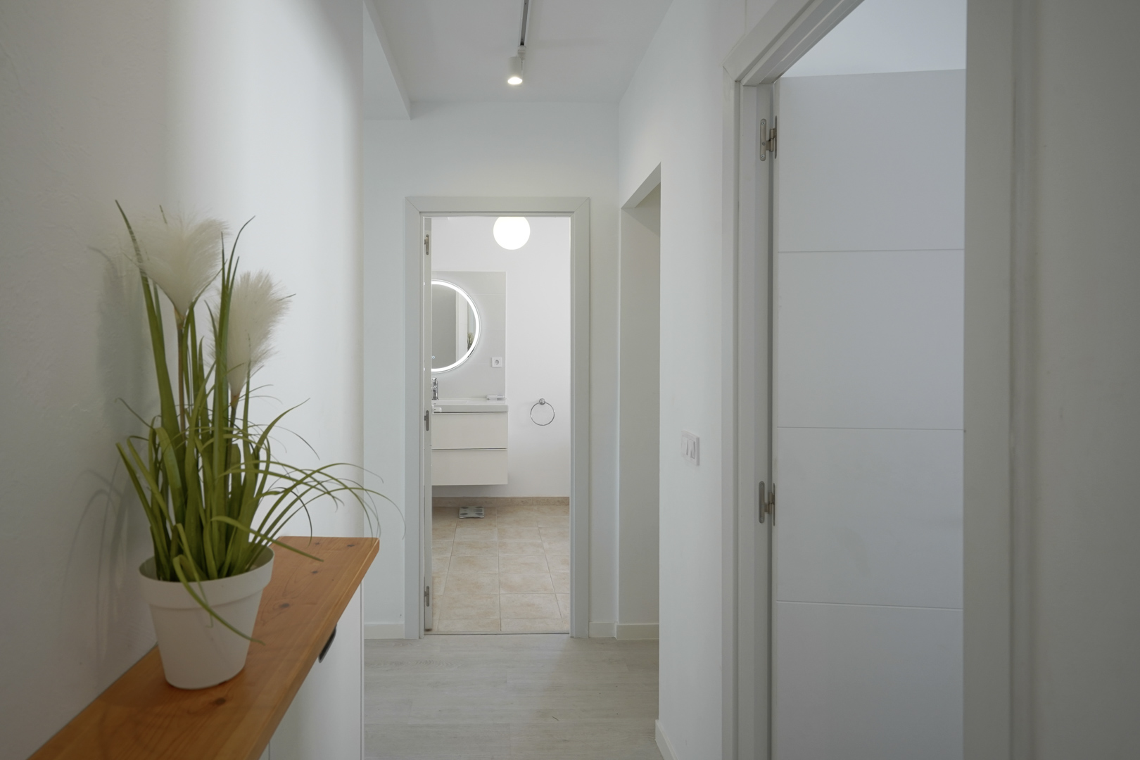 Heller Flur mit weißen Wänden und hellem Bodenbelag, der zu einem modernen Badezimmer mit hinterleuchtetem Rundspiegel und Hängeschrank führt.