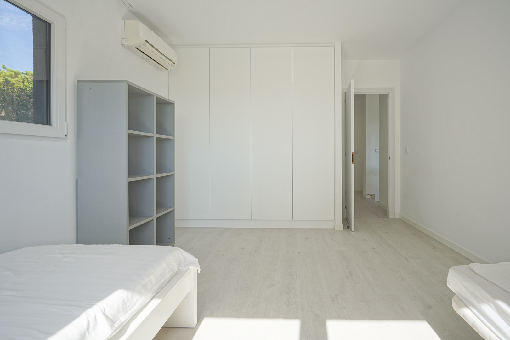 Helles Schlafzimmer mit deckenhohen weißen Einbauschränken, hellem Laminatboden, Wandklimaanlage und einem grauen Standregal in modernem Design.