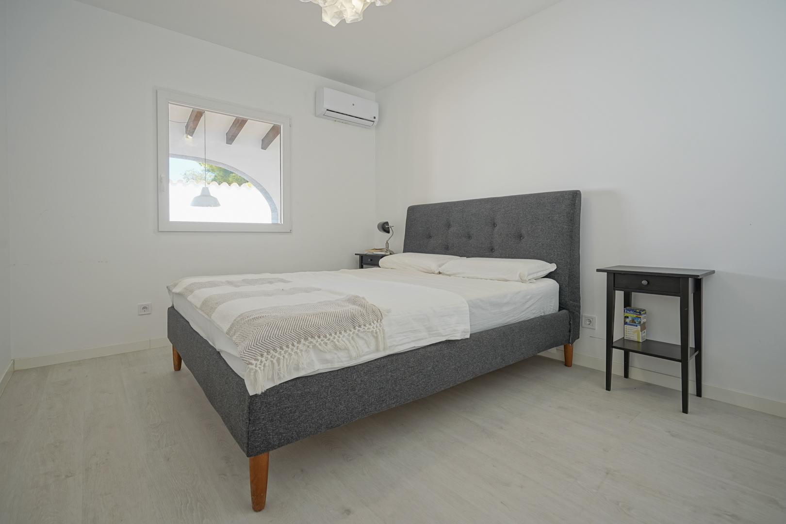 Helles Schlafzimmer mit grau gepolstertem Doppelbett, Klimaanlage und hellem Laminatboden. Das Fenster bietet einen Blick auf die überdachte Terrasse mit Holzbalken.