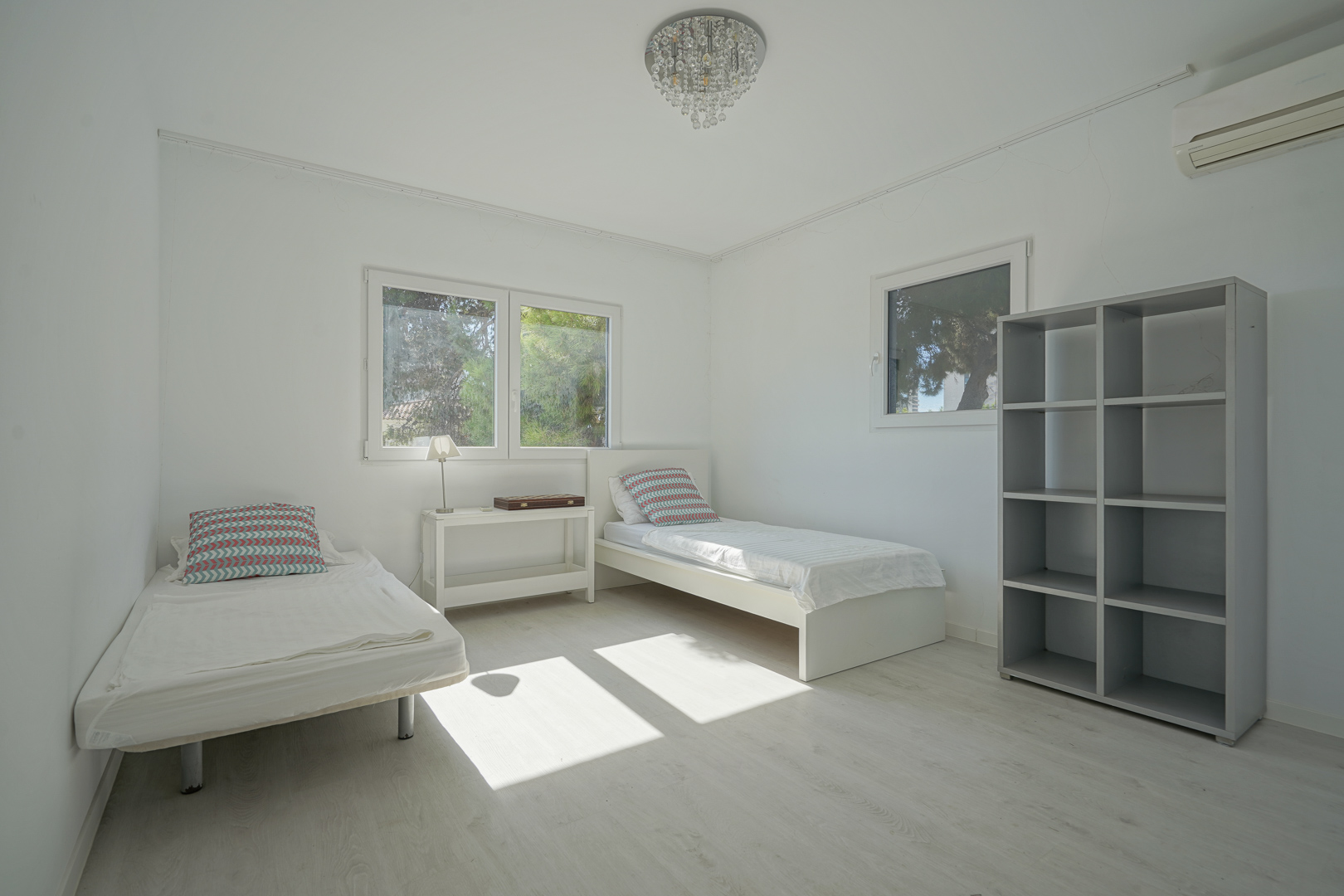 Helles Schlafzimmer mit zwei Einzelbetten, hellem Holzboden und grauem Regal. Ausgestattet mit Klimaanlage, Kristallleuchter und Fenstern mit Blick in den Garten.