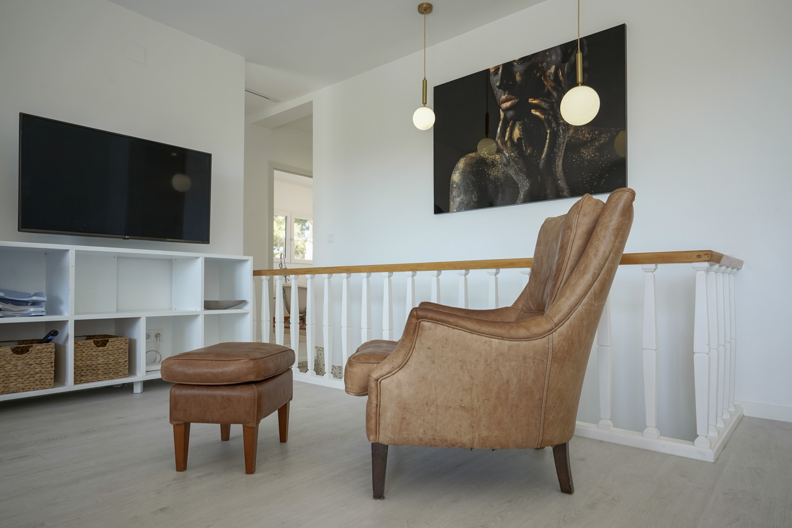 Leseecke mit Ledersessel und Hocker neben dem Treppengeländer. Ausgestattet mit Wandfernseher, weißem Regal und modernen Pendelleuchten.