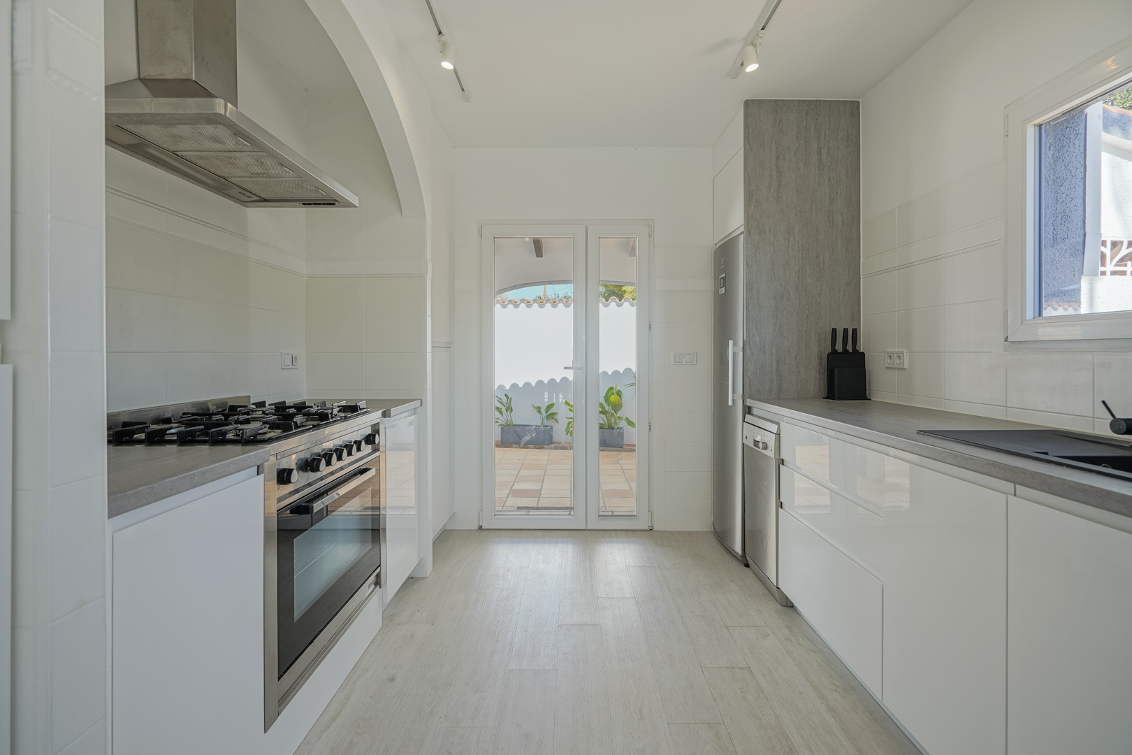 Moderne Küche mit glänzend weißen Schränken, grauen Arbeitsplatten und Edelstahlelementen. Ausgestattet mit Gasherd und direktem Zugang zur Außenterrasse.