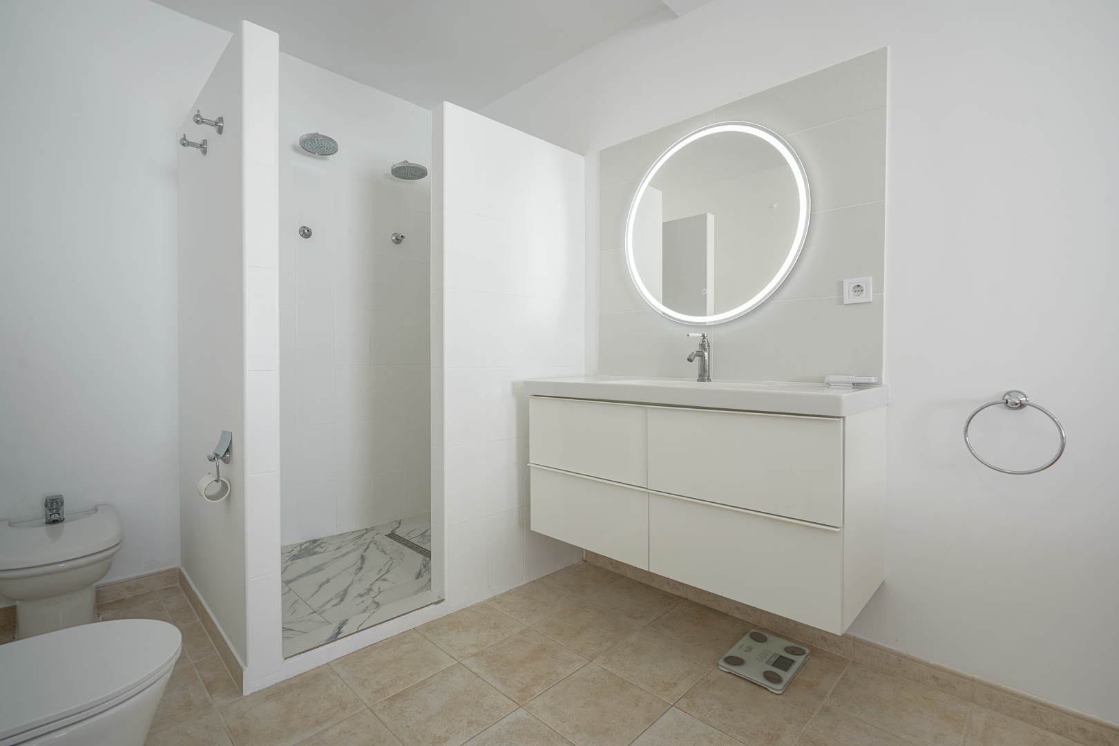 Modernes Badezimmer mit schwebendem weißem Waschtisch, hinterleuchtetem Rundspiegel und einer geräumigen Doppel-Walk-In-Dusche mit Boden in Marmoroptik.