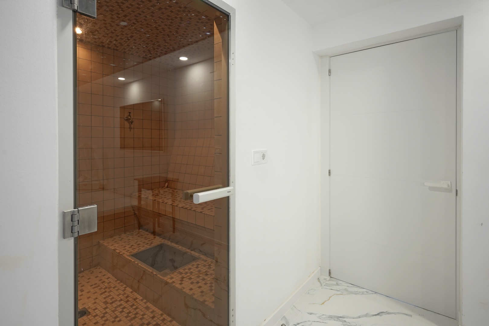 Private Sauna mit keramischer Mosaikverkleidung, Einbaustrahlern und Glastür, angrenzend an einen Raum mit edlem Marmorboden.