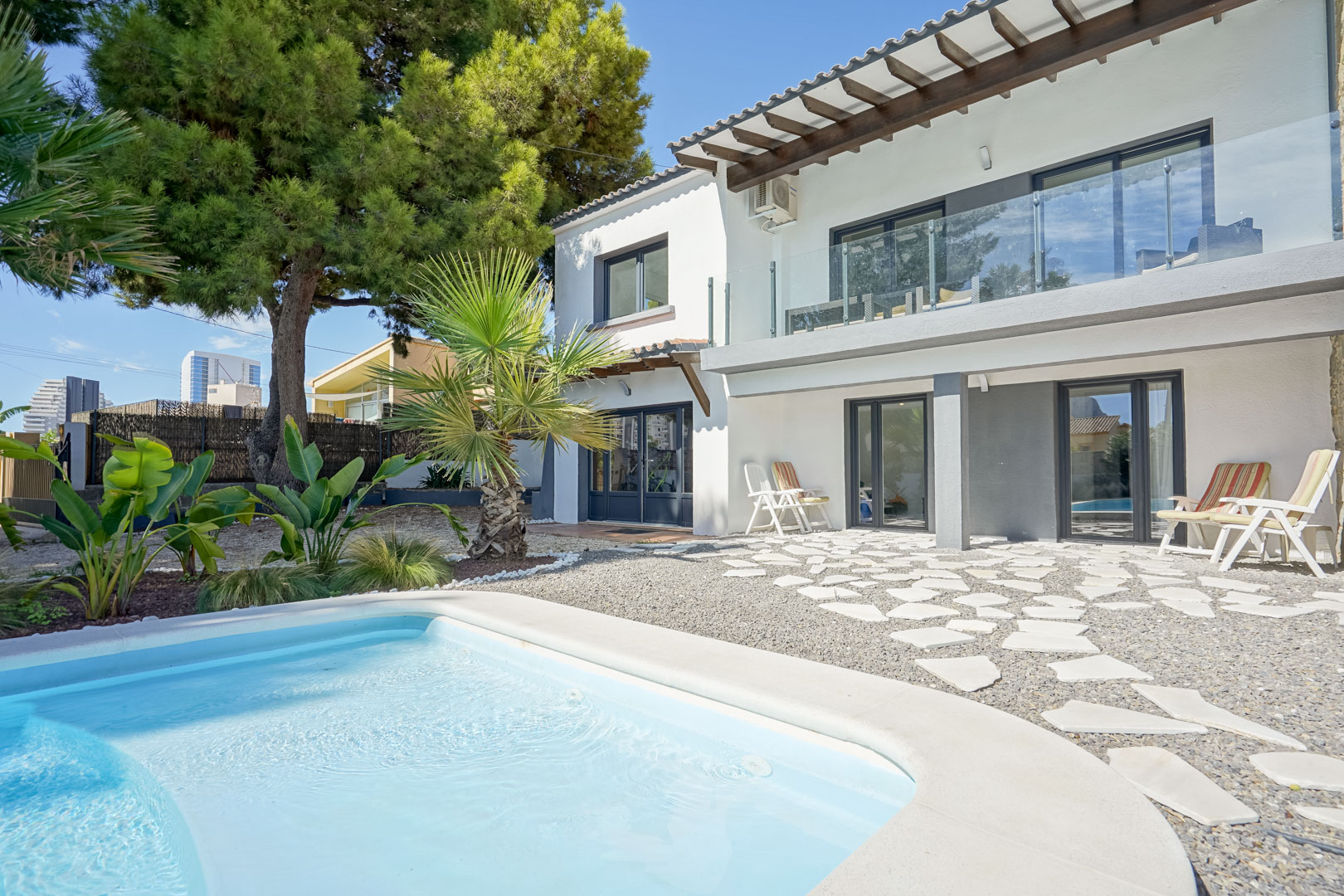 Rückansicht der Villa mit Pool, oberer Terrasse mit Glasgeländer und Holzbalken sowie einem Kiesgarten mit unregelmäßigen Trittsteinen.