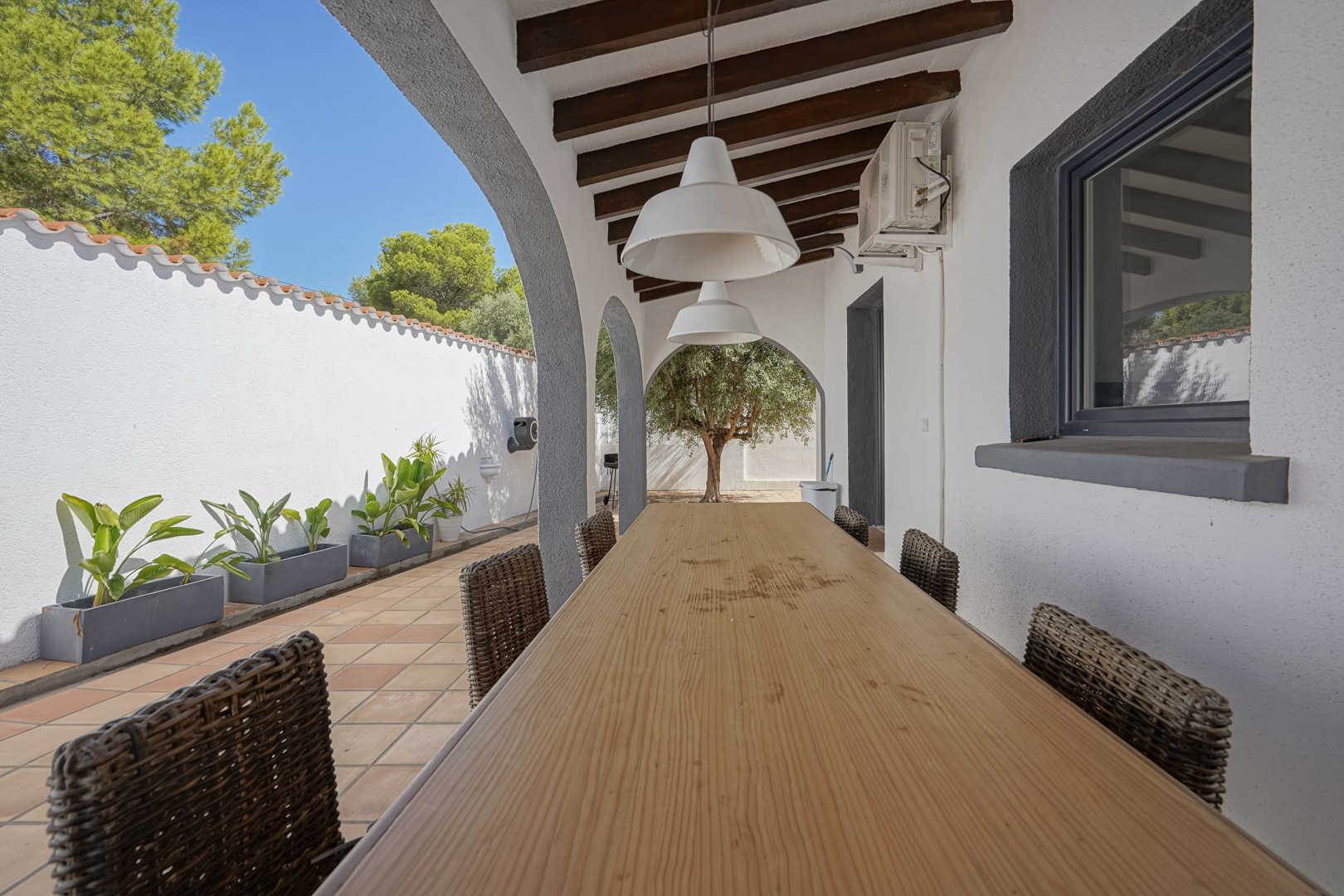 Überdachte Terrasse mit Holzbalkendecke, langem Esstisch und Korbstühlen. Die Rundbögen bieten einen Blick auf den mediterranen Garten mit Olivenbaum.