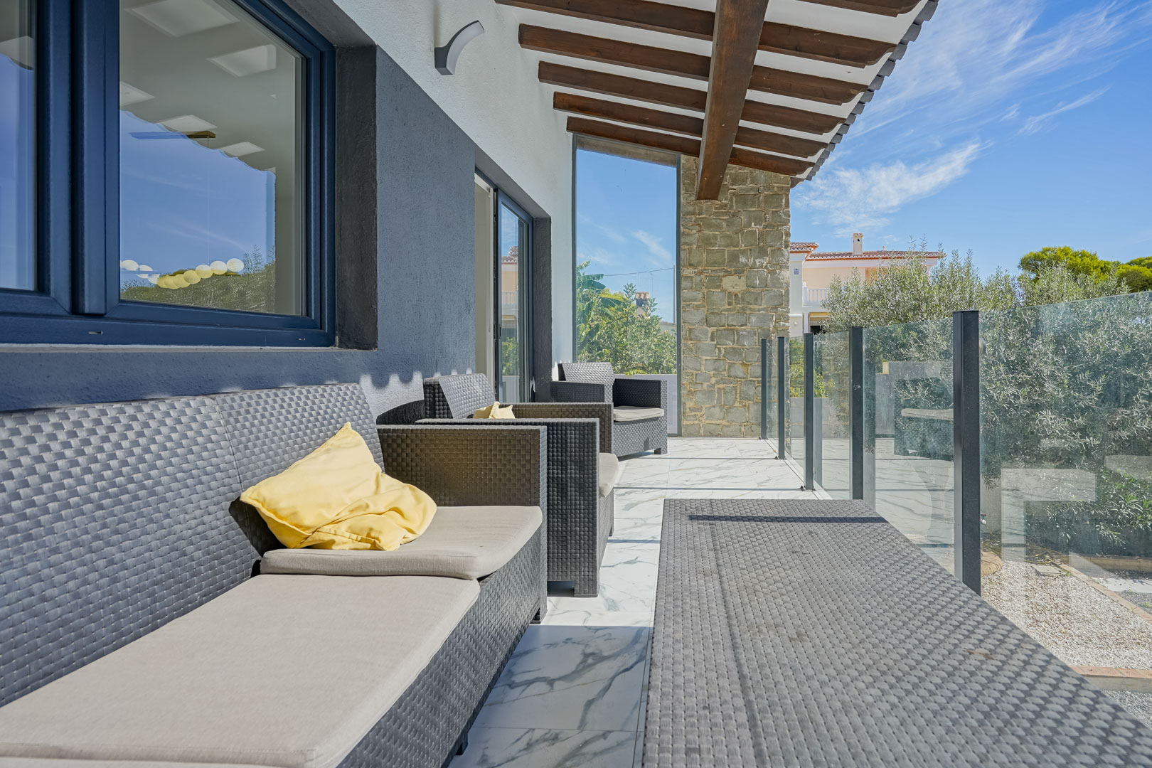 Überdachte Terrasse mit Holzbalkendecke, Polyrattan-Möbeln und Marmorboden. Die Glasbalustrade ermöglicht einen freien Blick auf die mediterrane Umgebung.