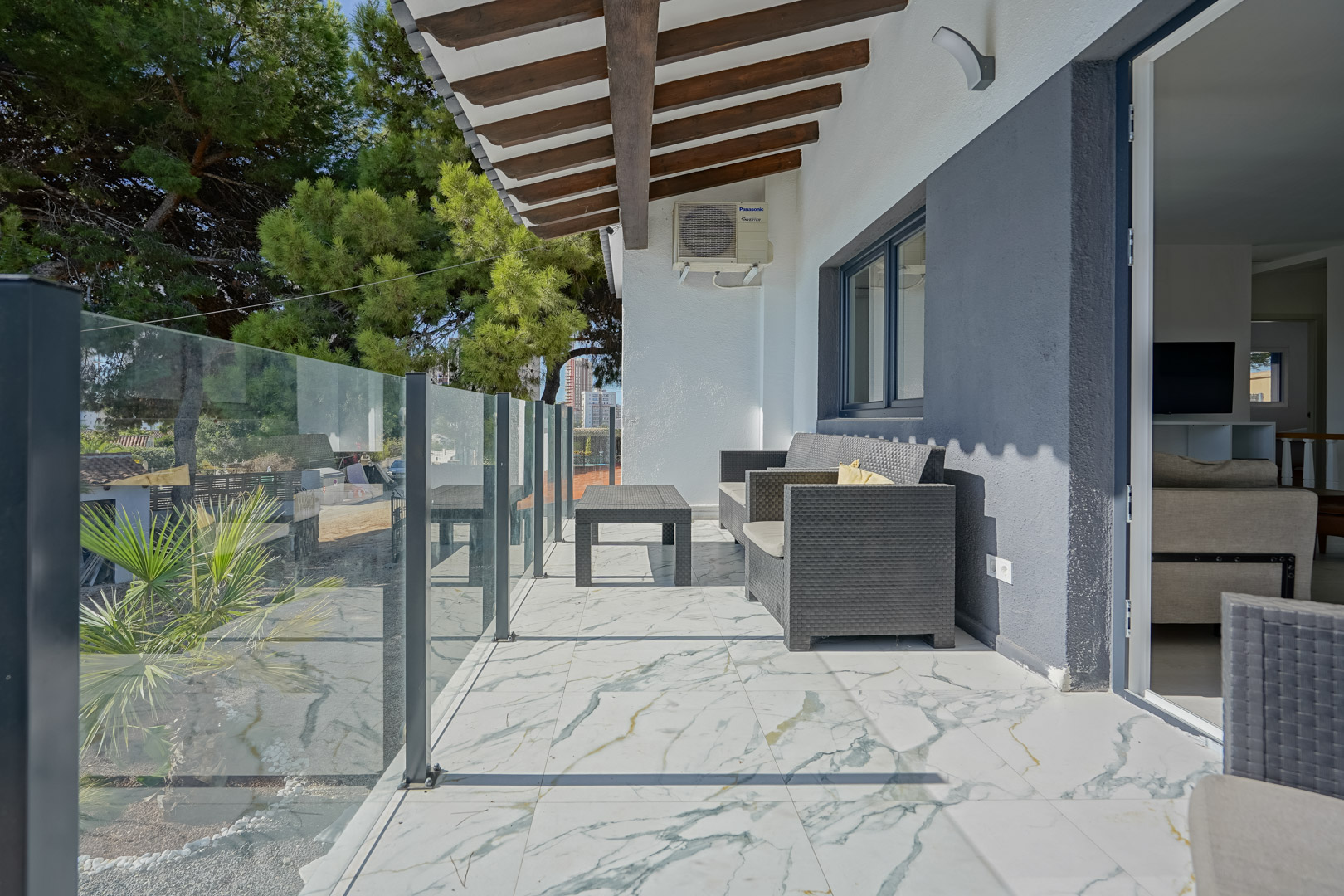Überdachte Terrasse mit marmorierten Bodenfliesen, Glasbalustrade und Holzbalkendecke. Ausgestattet mit grauen Rattanmöbeln und Blick auf die mediterrane Umgebung.