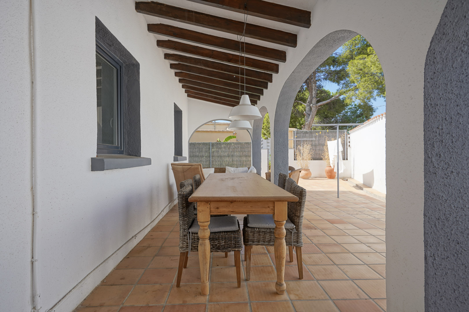 Überdachte Terrasse mit sichtbaren Holzbalken, mediterranen Rundbögen und Essbereich im Freien mit Holztisch und Korbstühlen auf Terrakottafliesen.