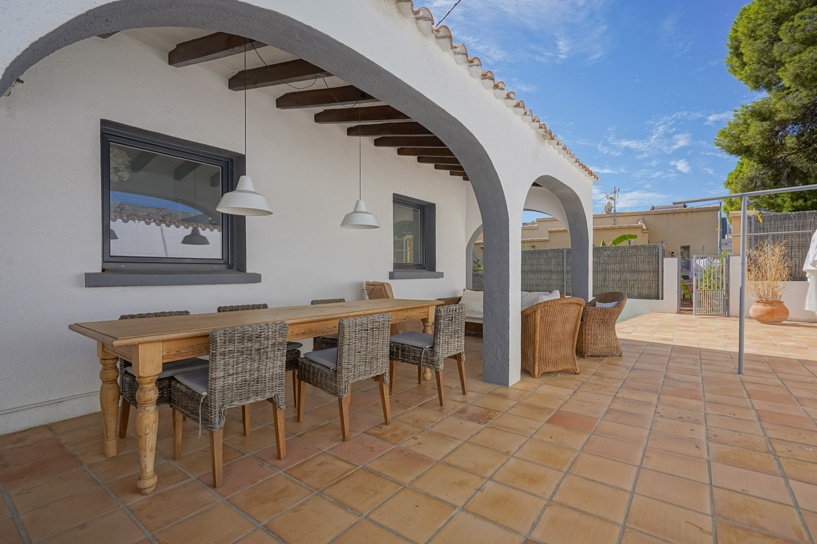 Überdachte Terrasse mit weißen Rundbögen, Holzbalkendecke und Terrakottafliesen. Ein Essbereich mit Holztisch und Korbstühlen lädt zum Verweilen ein.