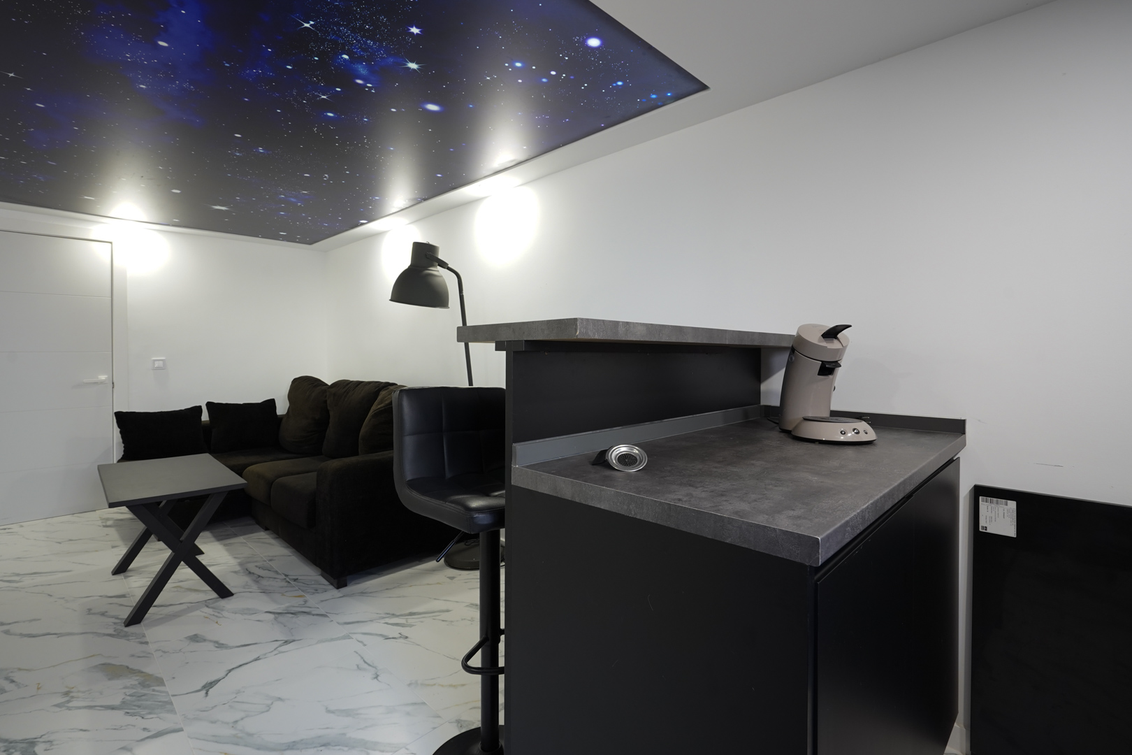 Wohnbereich mit Kaffeebar, dunklem Sofa und Marmoroptik-Fliesen, akzentuiert durch ein hinterleuchtetes Deckenpaneel mit Sternenhimmel-Design.