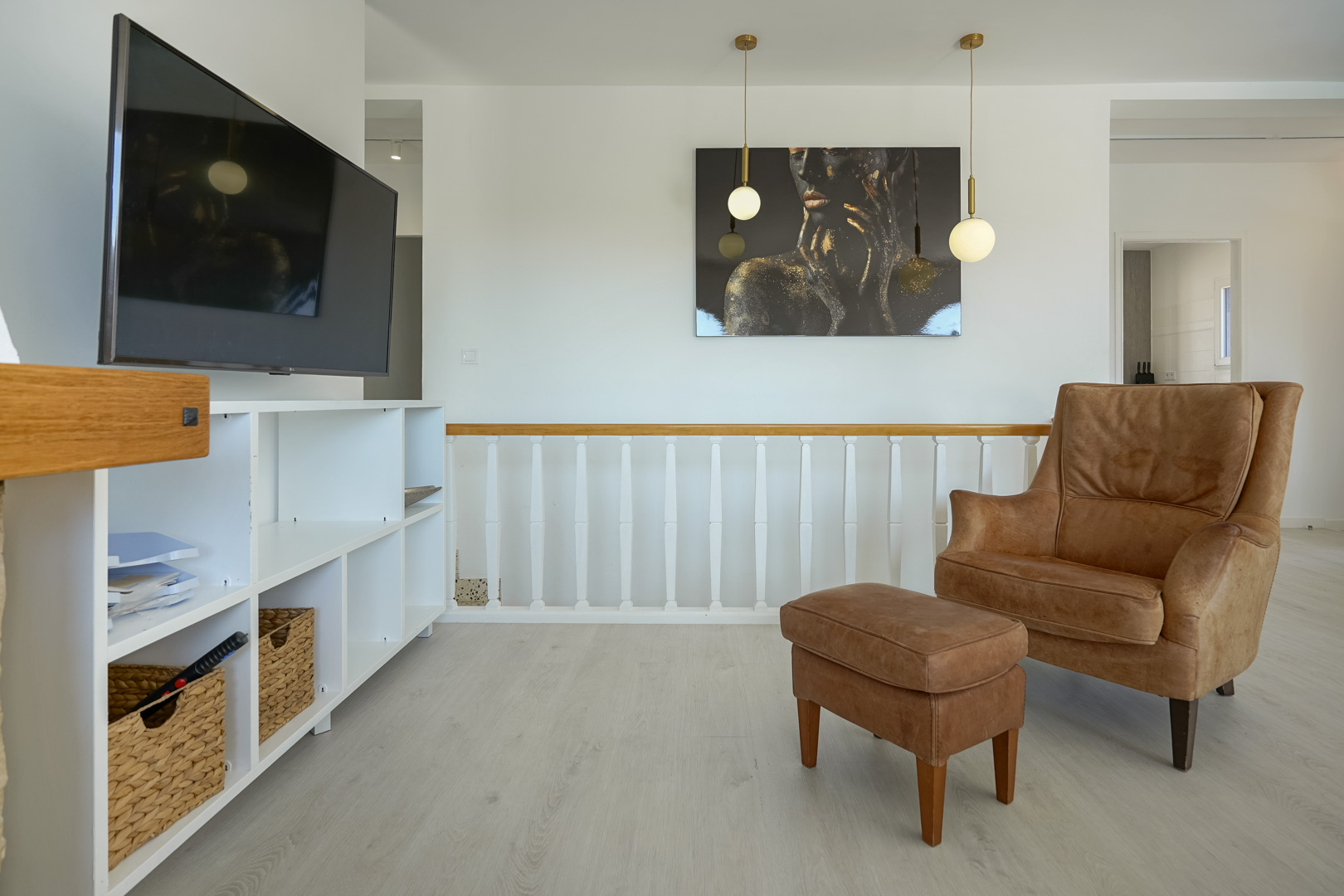 Wohnbereich mit Ledersessel und Hocker, weißem Regal und Holzgeländer. Moderne Pendelleuchten und heller Bodenbelag sorgen für ein zeitgemäßes Ambiente.