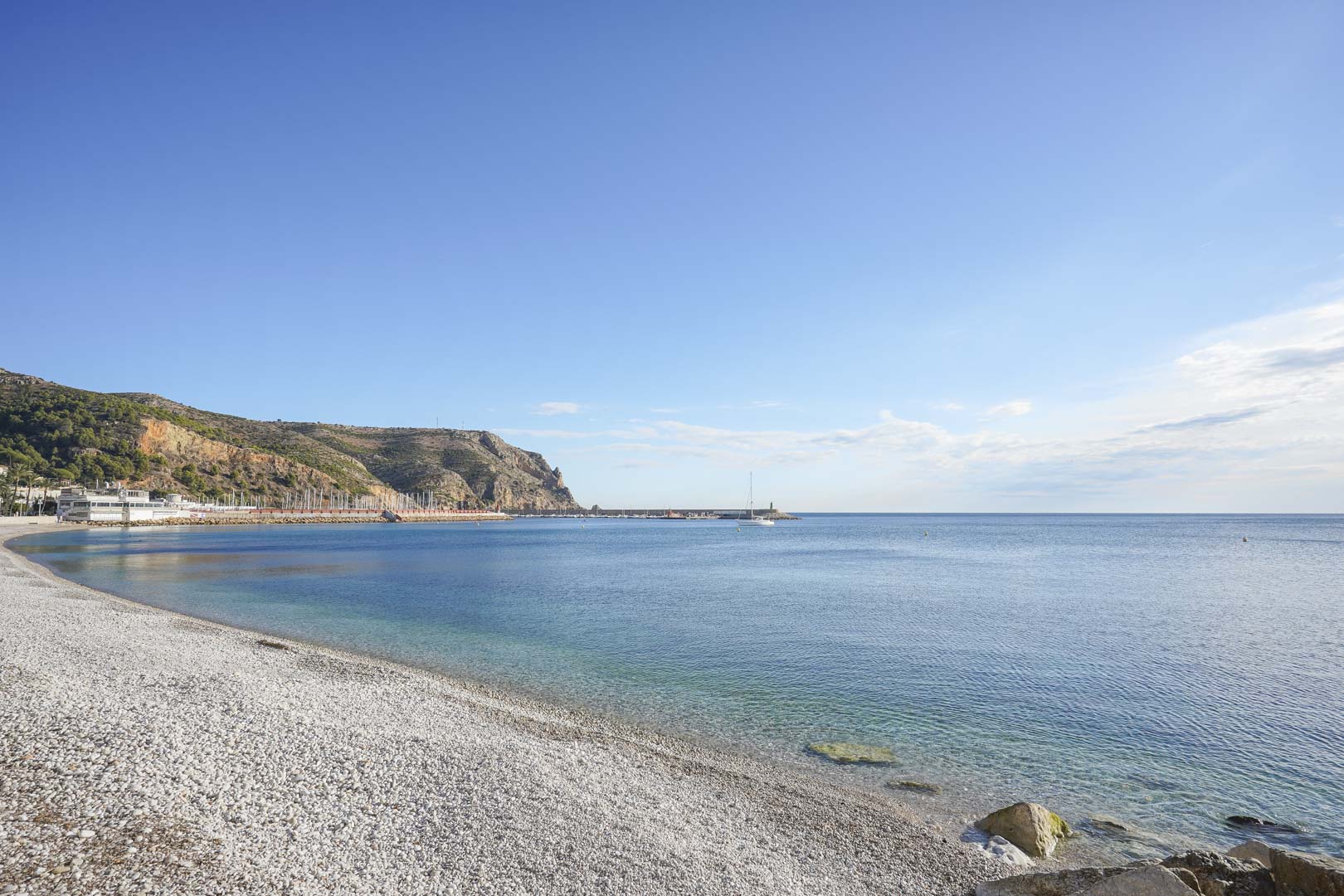 Panoramablick auf den Kieselstrand von Jávea mit klarem Mittelmeerwasser, dem Hafen und den markanten Klippen des Cabo de San Antonio.