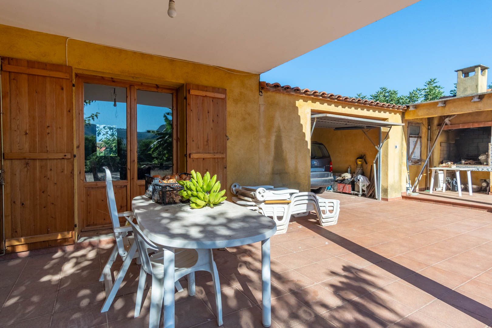 Außen-Terrasse mit Terrakotta-Fliesen, schattigem Essbereich und Zugang zum überdachten Parkplatz sowie zur traditionellen Außenküche mit Grill.