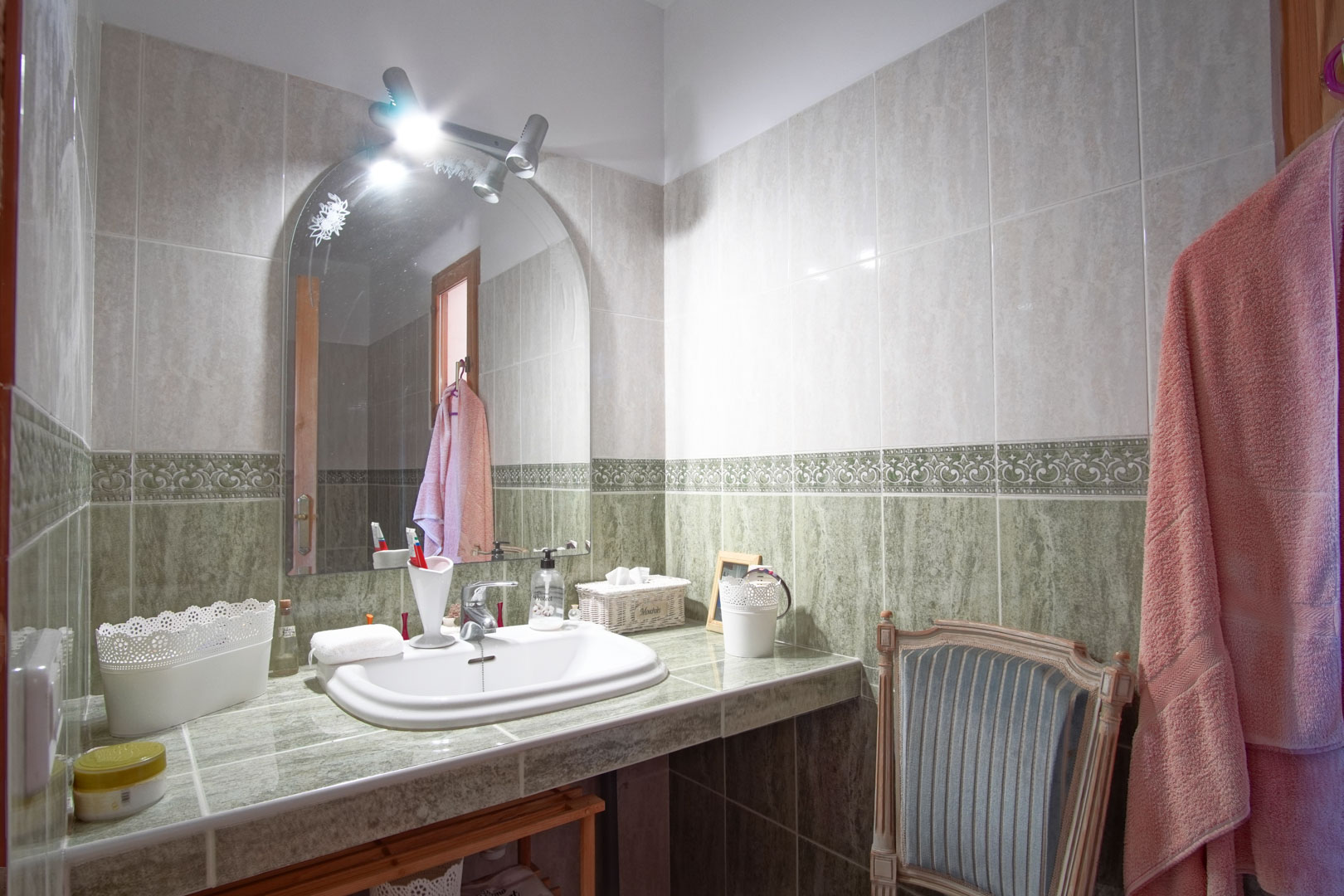 Badezimmer mit grün gefliester Arbeitsplatte, Einbauspülbecken, Rundbogenspiegel mit Spotbeleuchtung und dekorativer Bordüre an den Wänden.