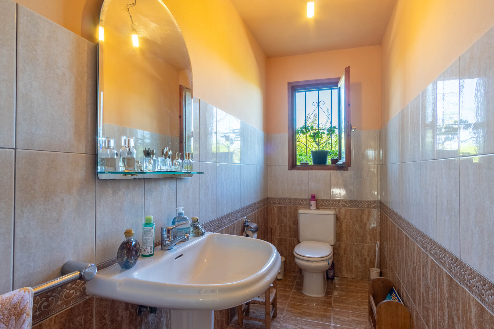 Badezimmer mit Wandfliesen in Erdtönen, Säulenwaschbecken, Bogenspiegel und einem Holzfenster mit Blick ins Grüne.