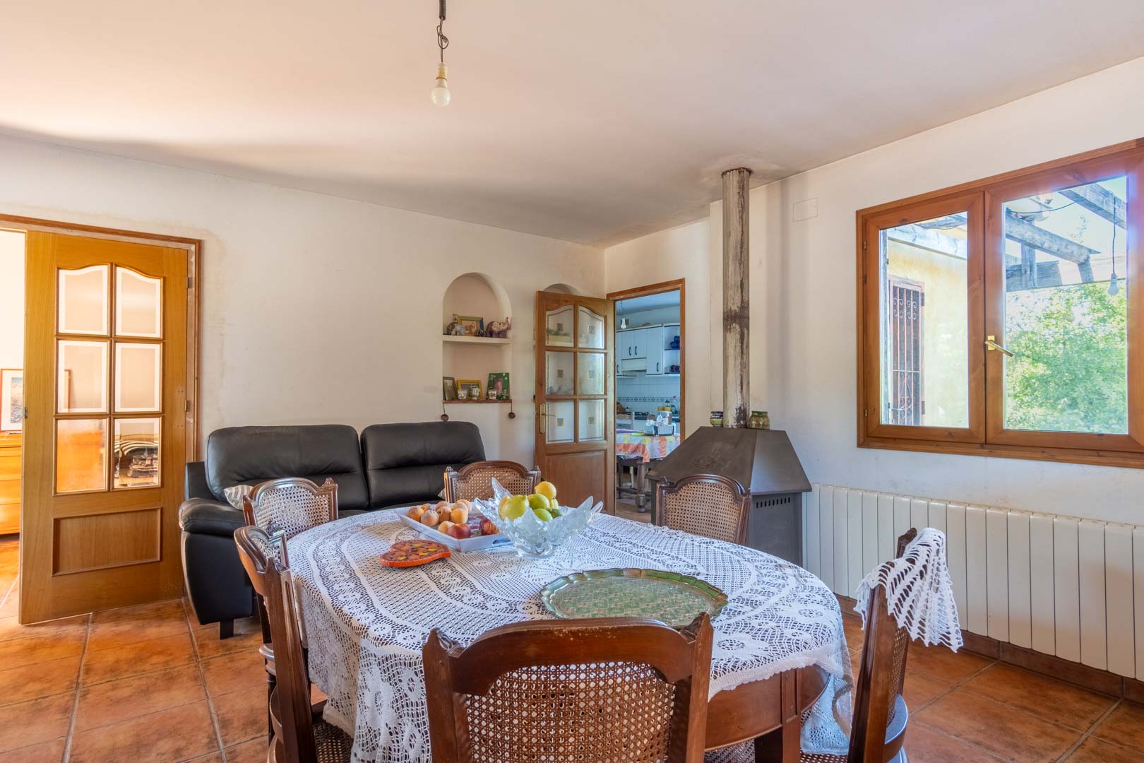 Essbereich mit Terrakottaboden, einem rustikalen Holzofen und klassischen Holzmöbeln. Große Fenster mit Holzrahmen sorgen für natürliches Licht in diesem funktionalen Raum.