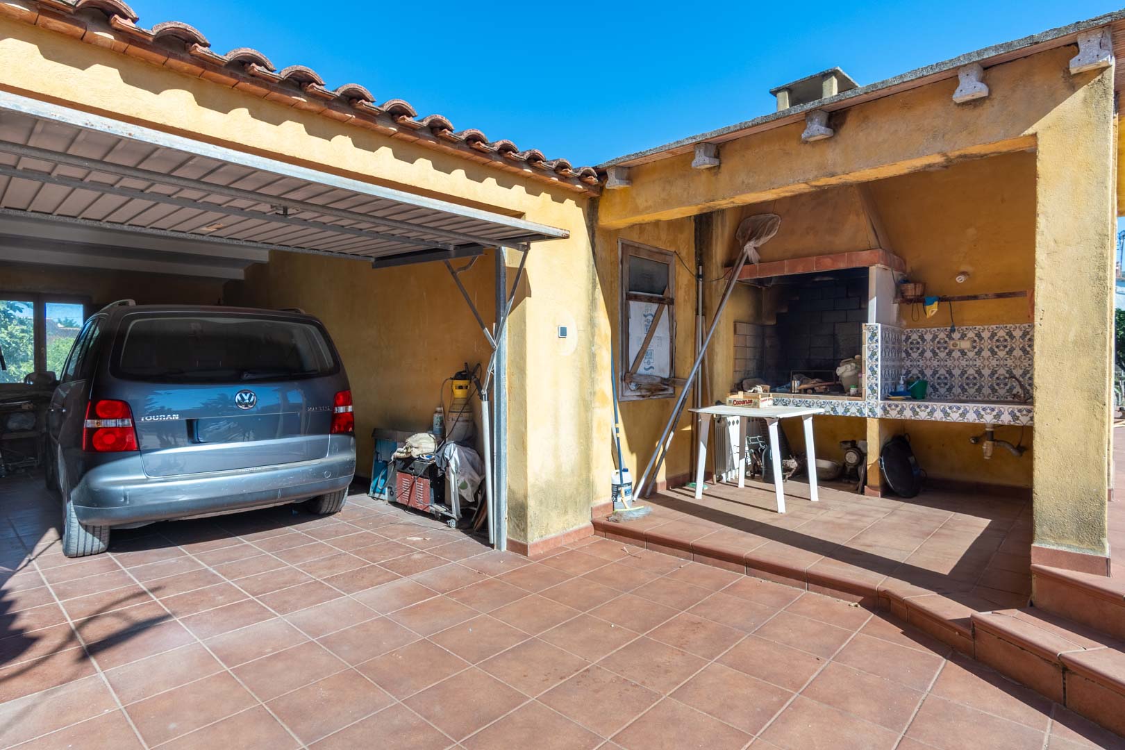 Funktionaler Außenbereich mit überdachtem Parkplatz und traditionellem gemauertem Grillplatz mit gefliestem Arbeitsbereich.