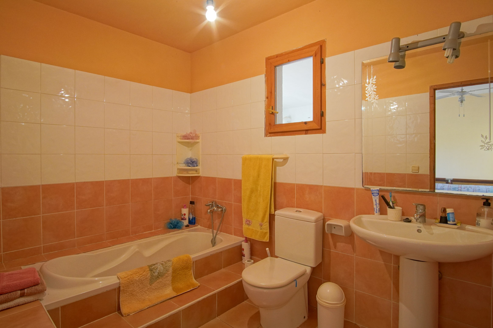 Funktionales Badezimmer mit lachsfarbenen und weißen Fliesen, Einbaubadewanne mit Einstiegsstufe, Standwaschbecken und Fenster mit Naturholzrahmen.