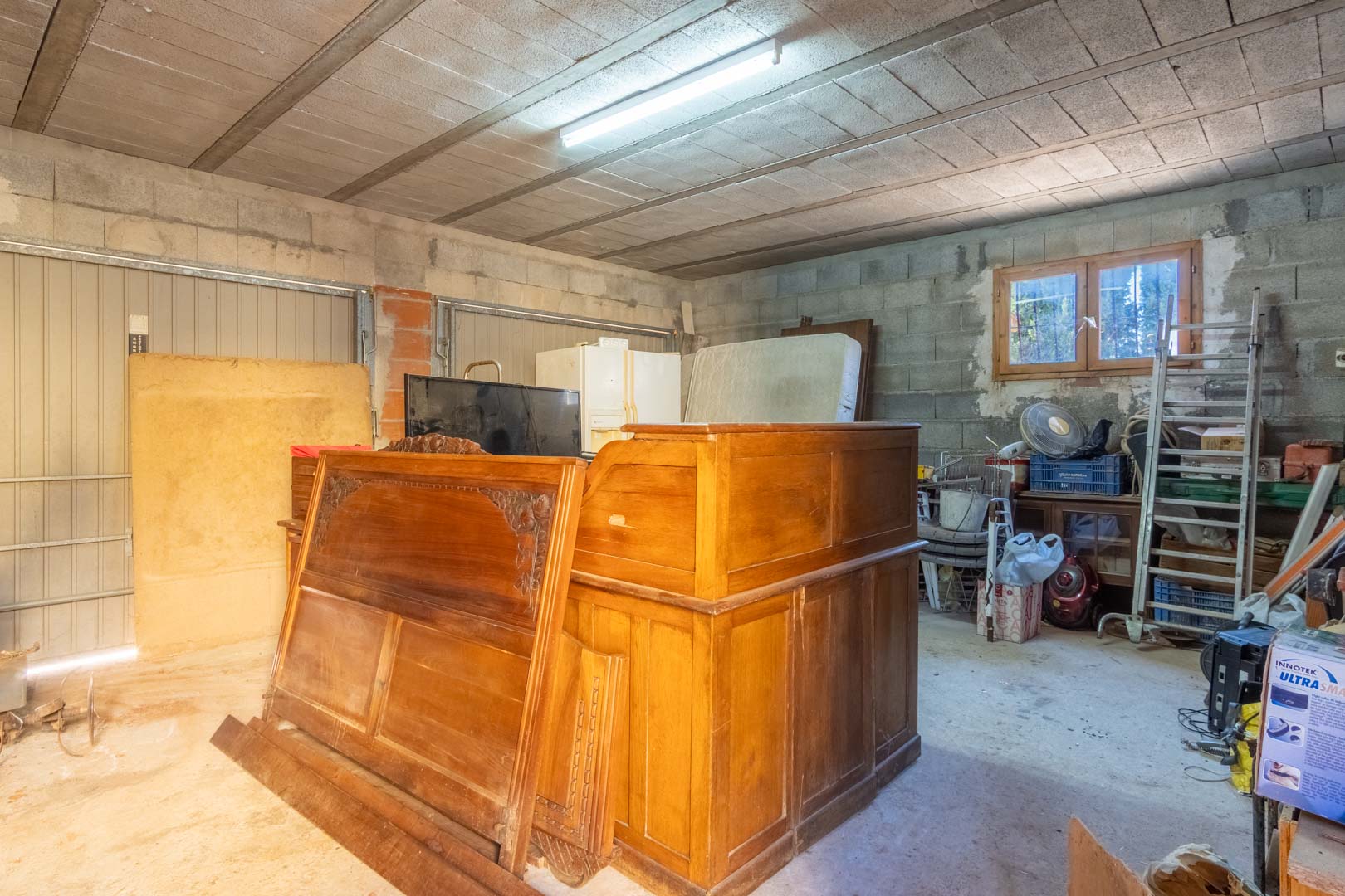 Geräumiger Lagerraum oder Garage mit Betonblockwänden, Balkendecke, Leuchtstoffröhren und einem Holzfenster, das für Tageslicht sorgt.
