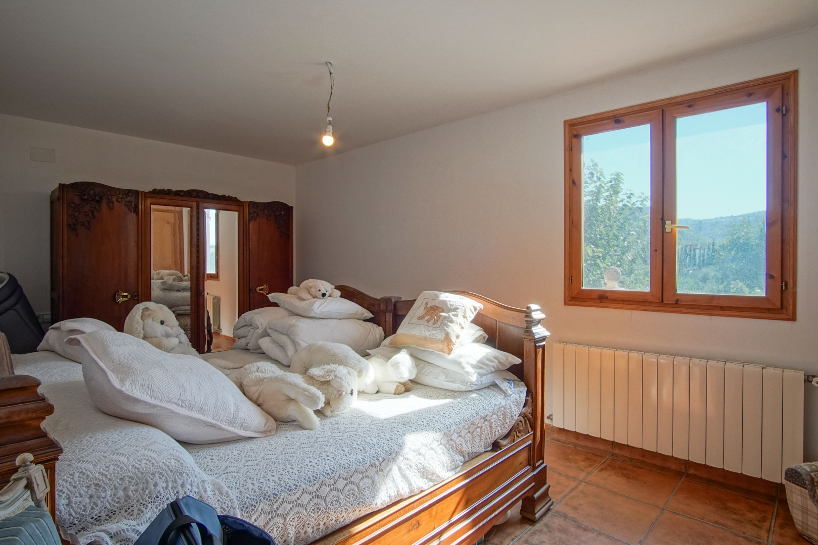 Schlafzimmer mit geschnitzten Holzmöbeln, Spiegelschrank, Terrakottaboden und Fenster mit Bergblick sowie Heizkörper.