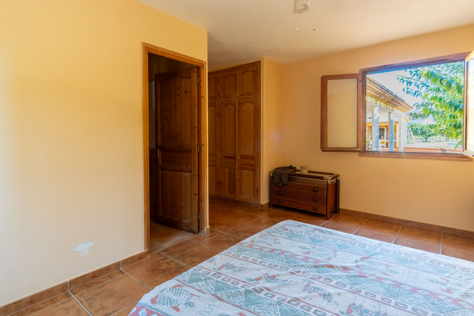 Schlafzimmer mit rustikalen Terrakottafliesen, Einbauschränken aus Holz und Fenster mit Blick auf die Veranda und den Garten.