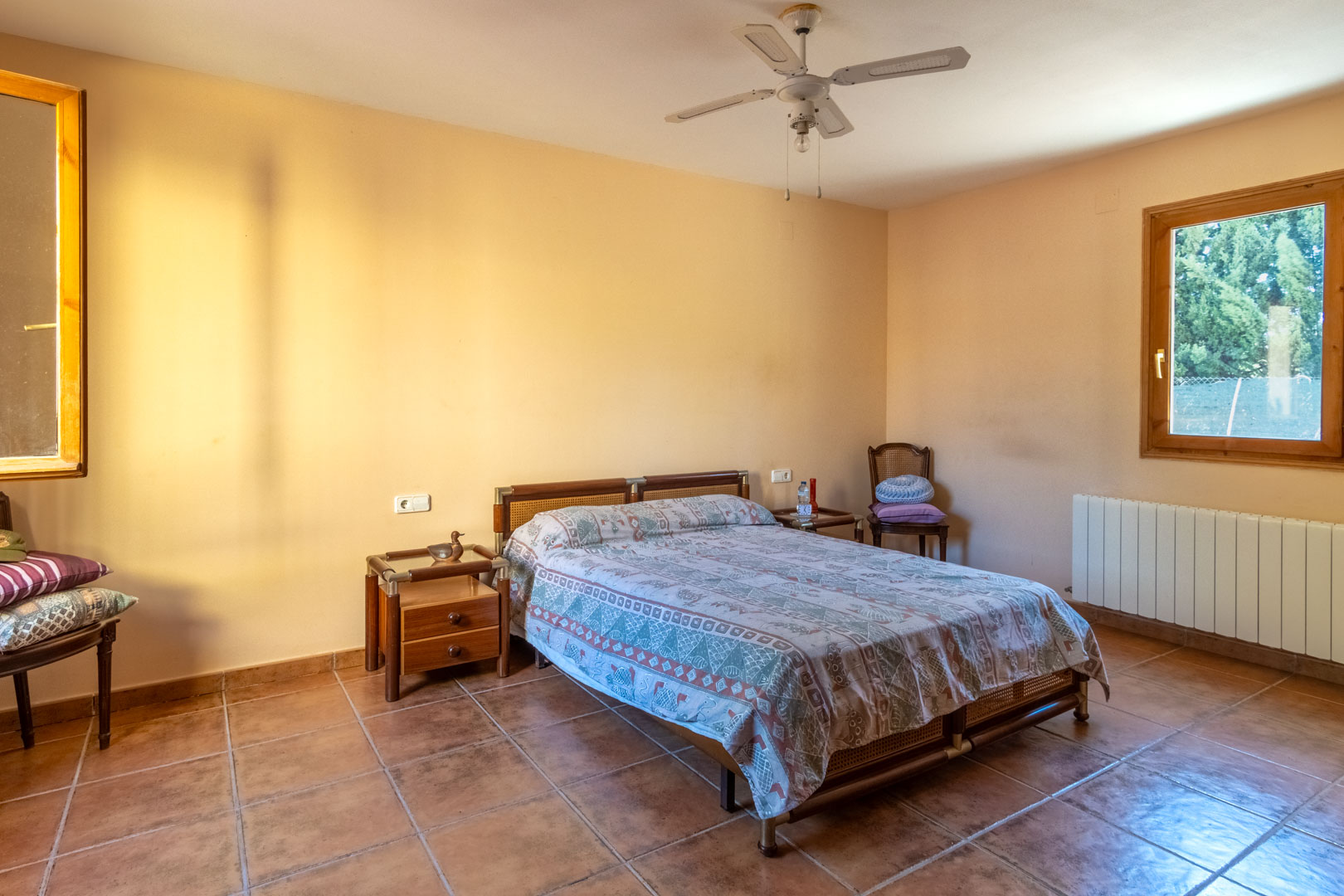 Schlafzimmer mit Terrakottaboden, warmen Wandfarben und Deckenventilator. Ausgestattet mit Holzfenstern, einem Heizkörper und rustikalen Nachttischen aus Holz und Glas.