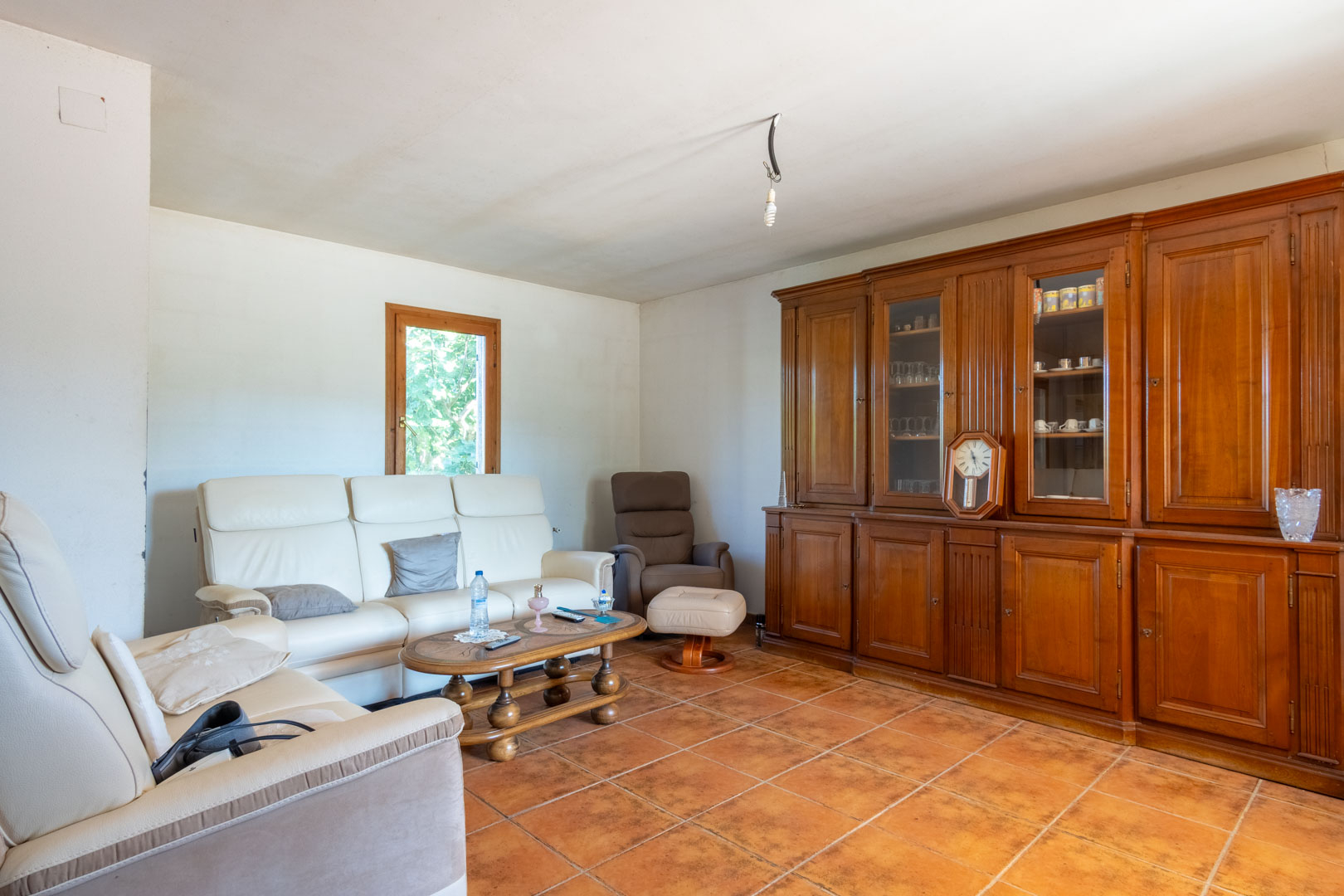 Wohnbereich mit rustikalem Fliesenboden, weißen Ledersofas und einer großen Schrankwand aus Massivholz mit Vitrineneinsätzen.