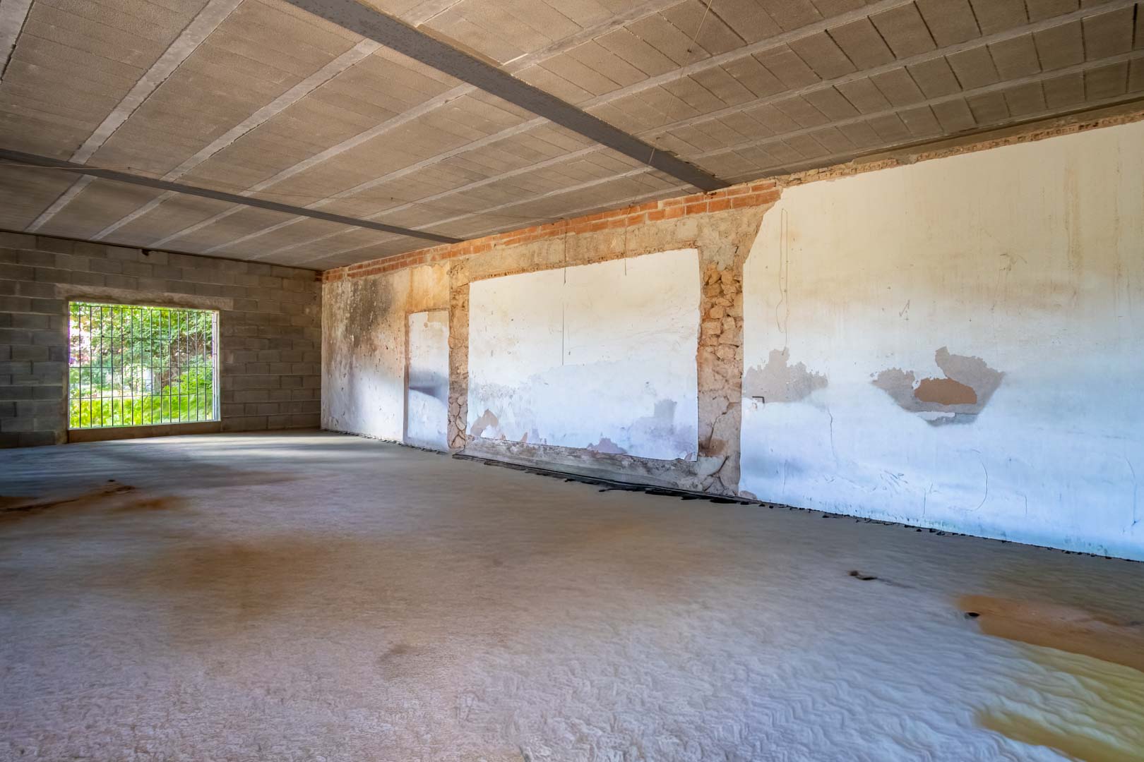 Offener Innenraum im Rohbauzustand mit Betonboden, sichtbaren Deckenbalken und Wänden aus einer Kombination von Naturstein und Ziegelmauerwerk.