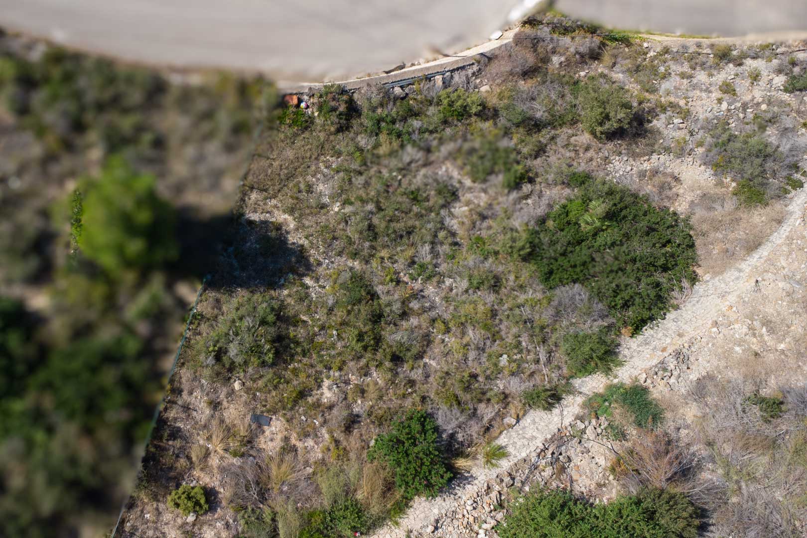 Luftaufnahme des abfallenden Baugrundstücks mit typisch mediterraner Vegetation, felsigem Untergrund und einem angrenzenden Steinpfad.