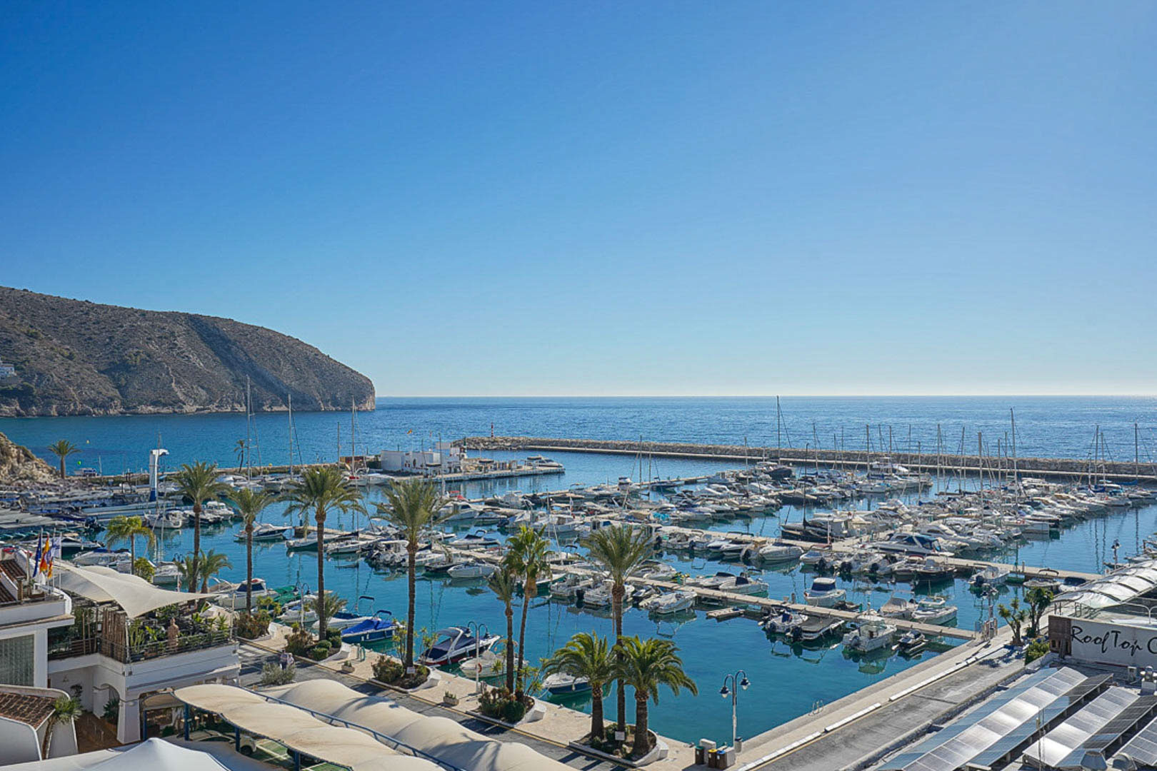 Panoramablick auf den Yachthafen von Moraira mit zahlreichen Booten, Palmen und der markanten Landzunge im Hintergrund.