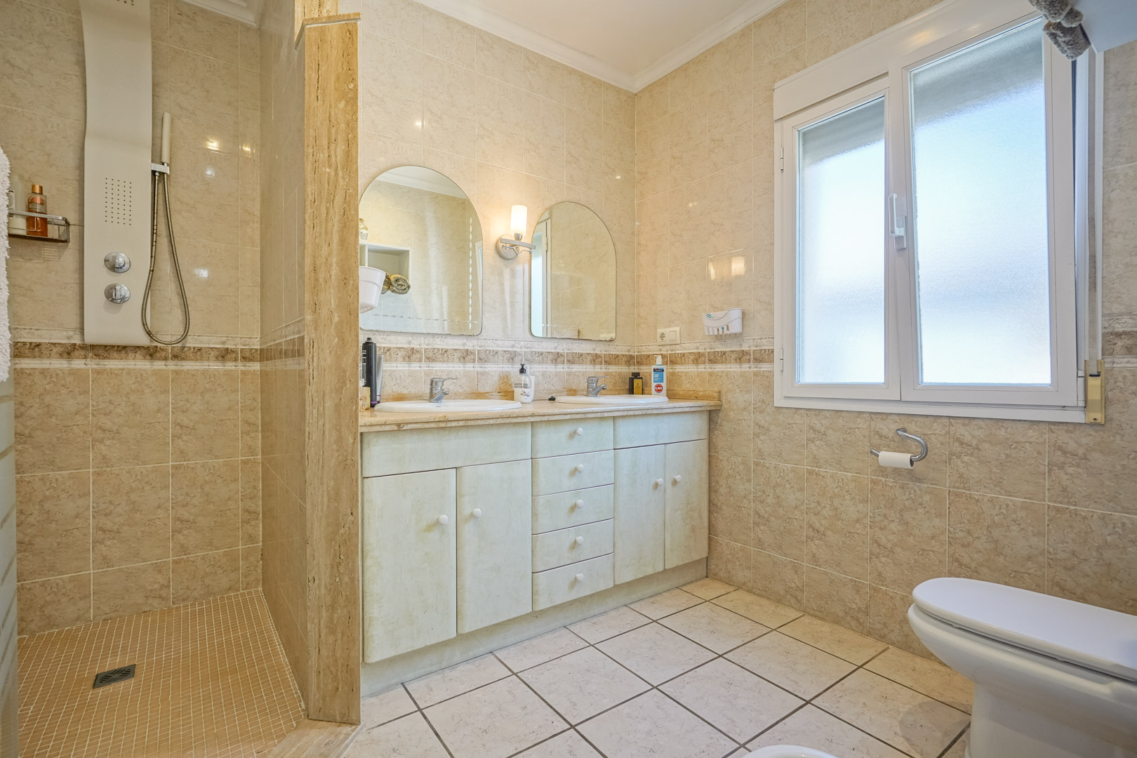 Badezimmer mit ebenerdiger Dusche, Waschtisch mit Doppelwaschbecken und cremefarbenen Fliesen.