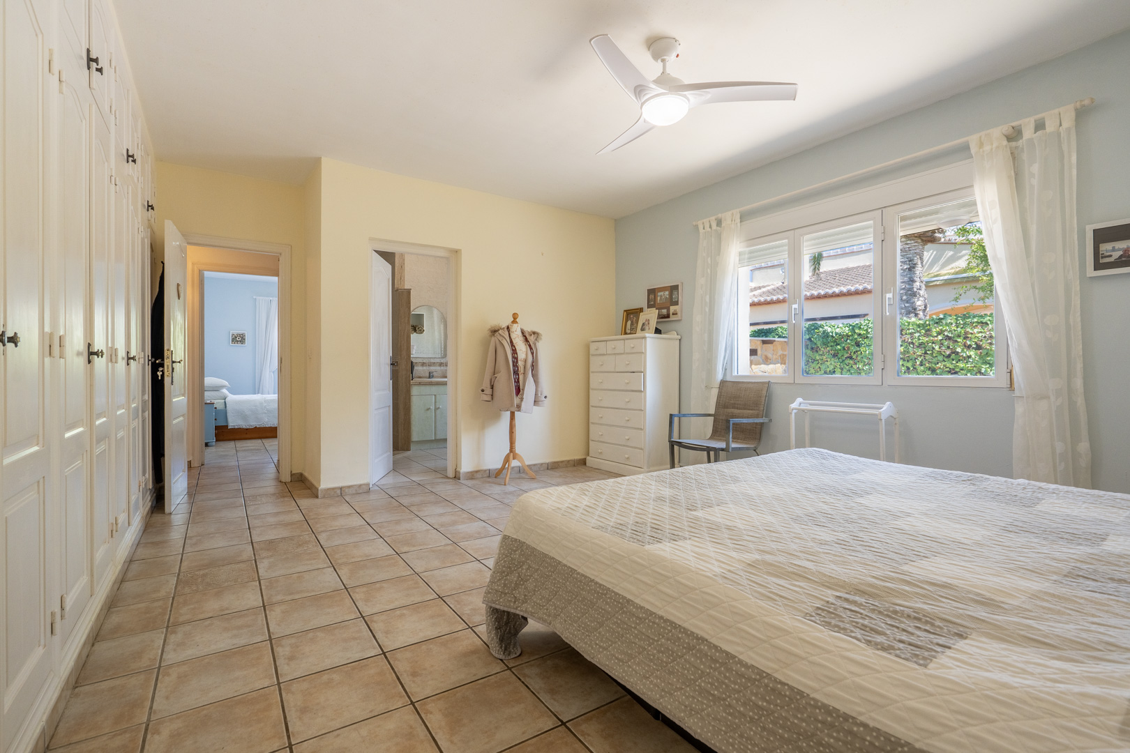 Helles Schlafzimmer mit Einbauschränken, Deckenventilator und Bad en Suite. Cremefarbene Fliesen.