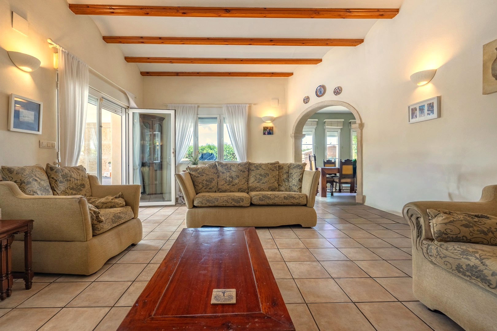 Helles Wohnzimmer mit Holzbalken, gemusterten Sofas und Fliesenboden. Zugang zum Esszimmer.