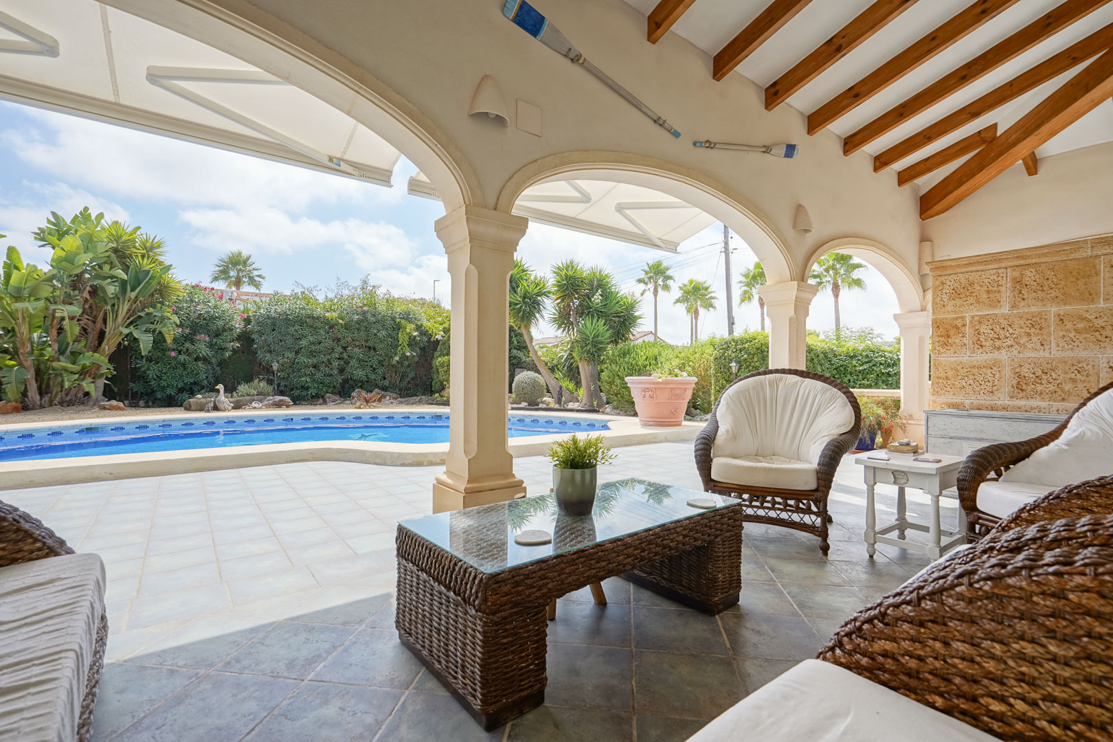 Überdachte Terrasse mit Blick auf den Pool. Mit Rattanmöbeln und einem Couchtisch mit Glasplatte.