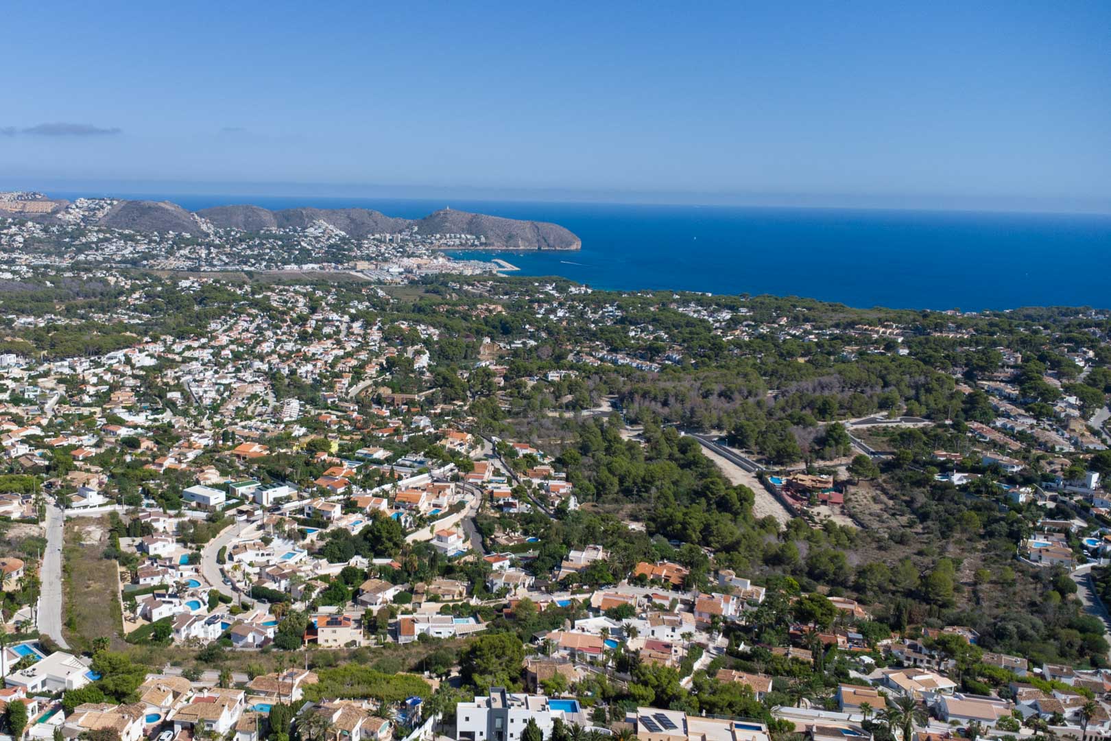 Panorama-Luftaufnahme von Moraira mit Blick auf die Mittelmeerküste, den Hafen und das Cap d'Or, eingebettet in Wohngebiete und Pinienwälder.