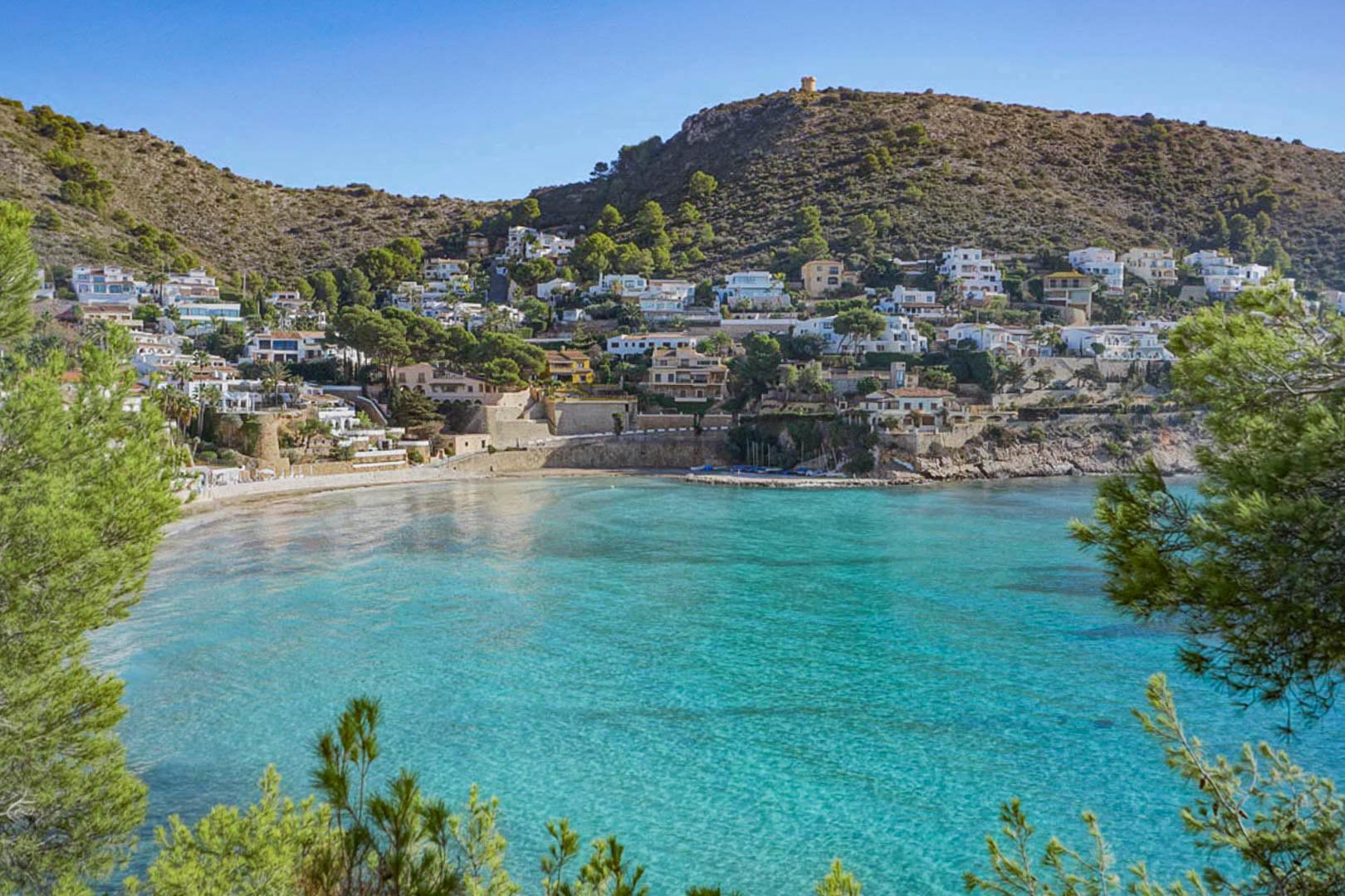 Panoramablick auf die Bucht von El Portet in Moraira mit türkisfarbenem Wasser, Villen am Hang und dem historischen Wachturm auf dem Gipfel des Berges.