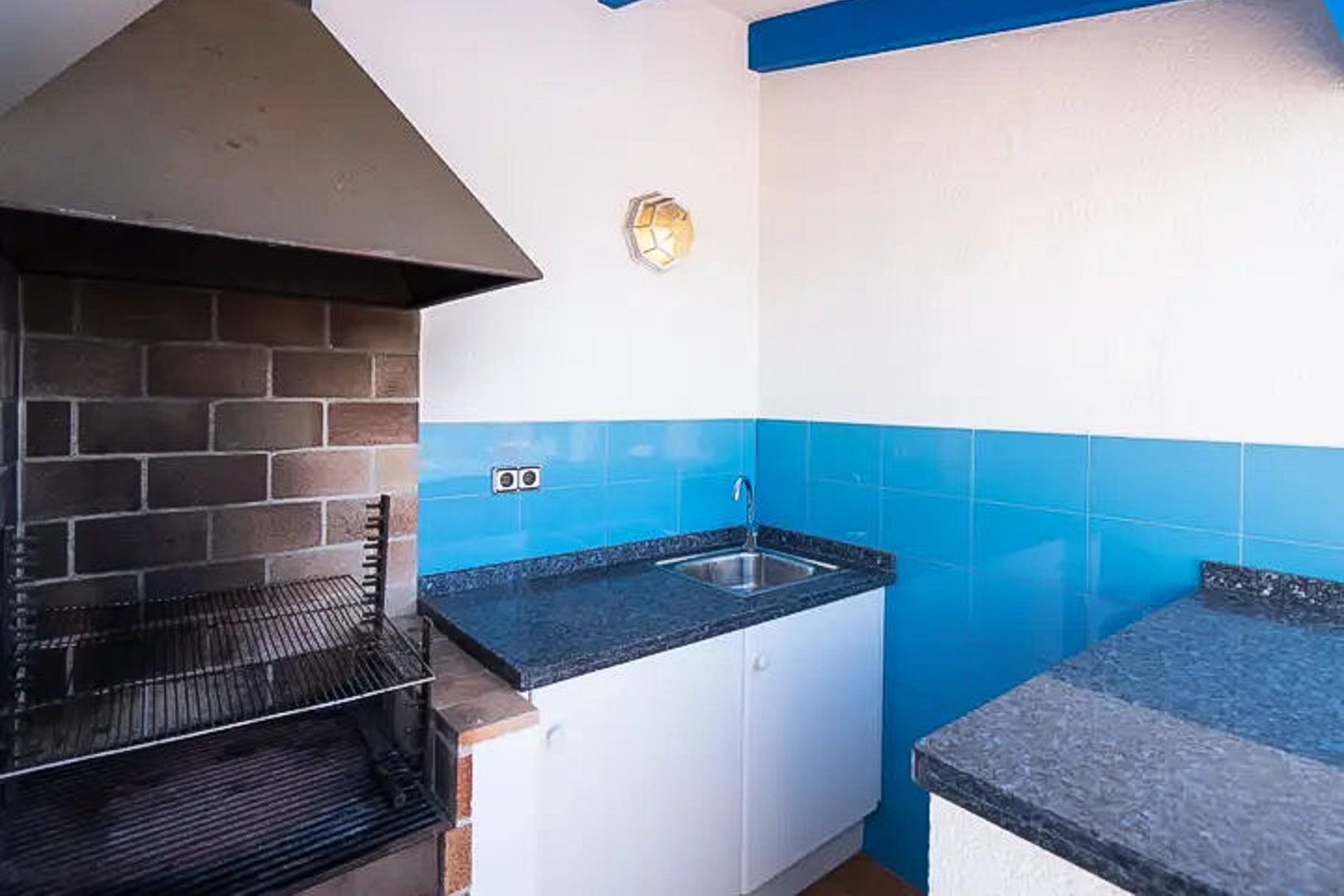 Außen-Grillbereich mit Metallhaube, dunkler Granitarbeitsplatte, Edelstahlspüle und mediterranen blauen Fliesen.