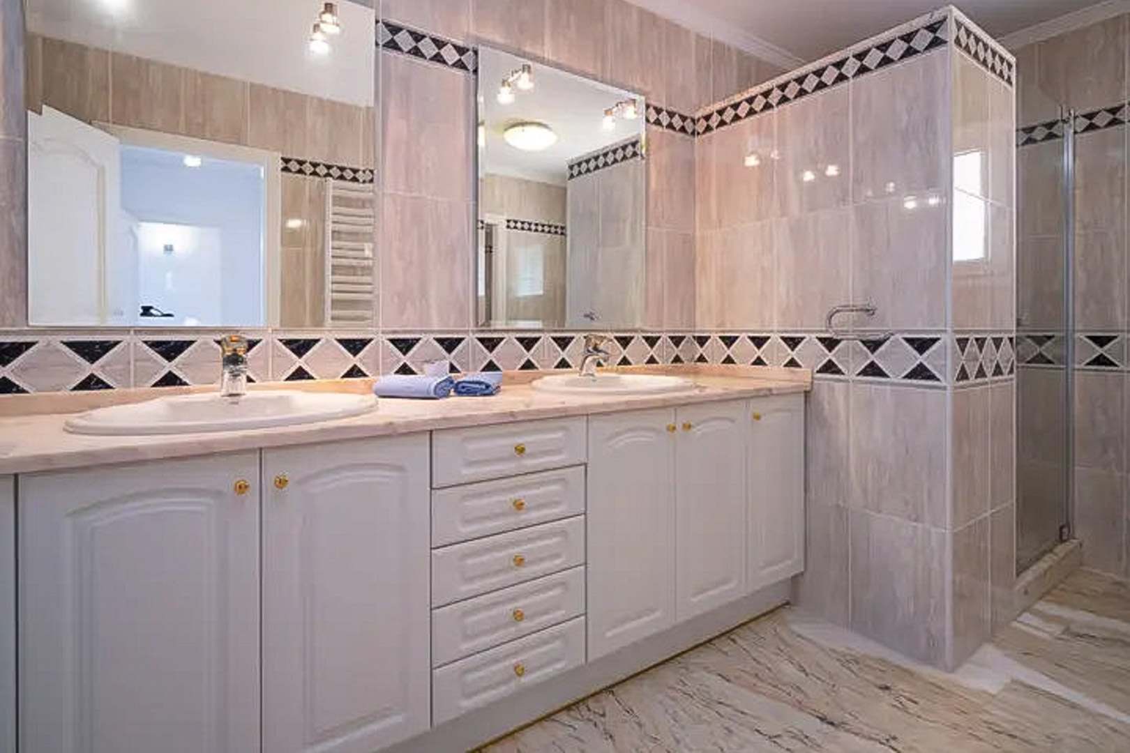 Badezimmer mit Doppelwaschtisch, weißen Schränken und Marmorplatte. Die Wände sind mit beigen Fliesen und einer dekorativen Rautenbordüre gestaltet.