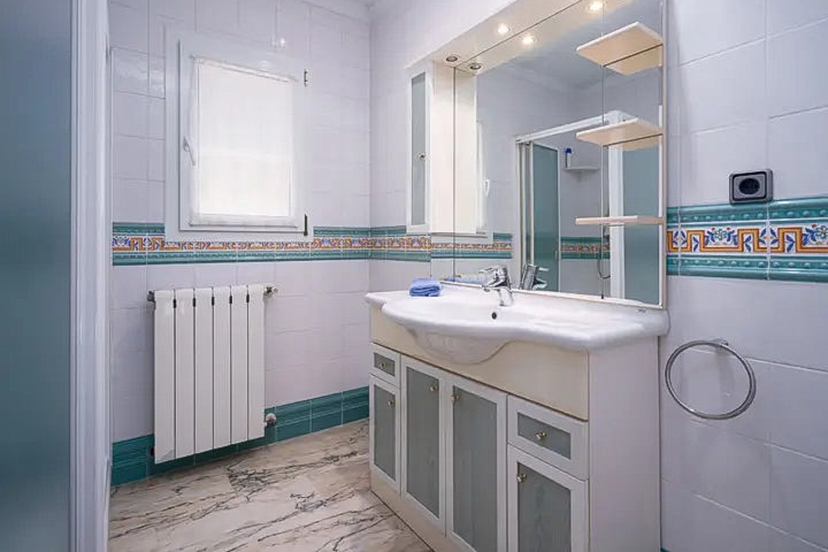 Badezimmer mit weißen Fliesen und mediterraner Bordüre, Waschtisch mit Milchglastüren, großem beleuchtetem Spiegel und Boden in Marmoroptik.
