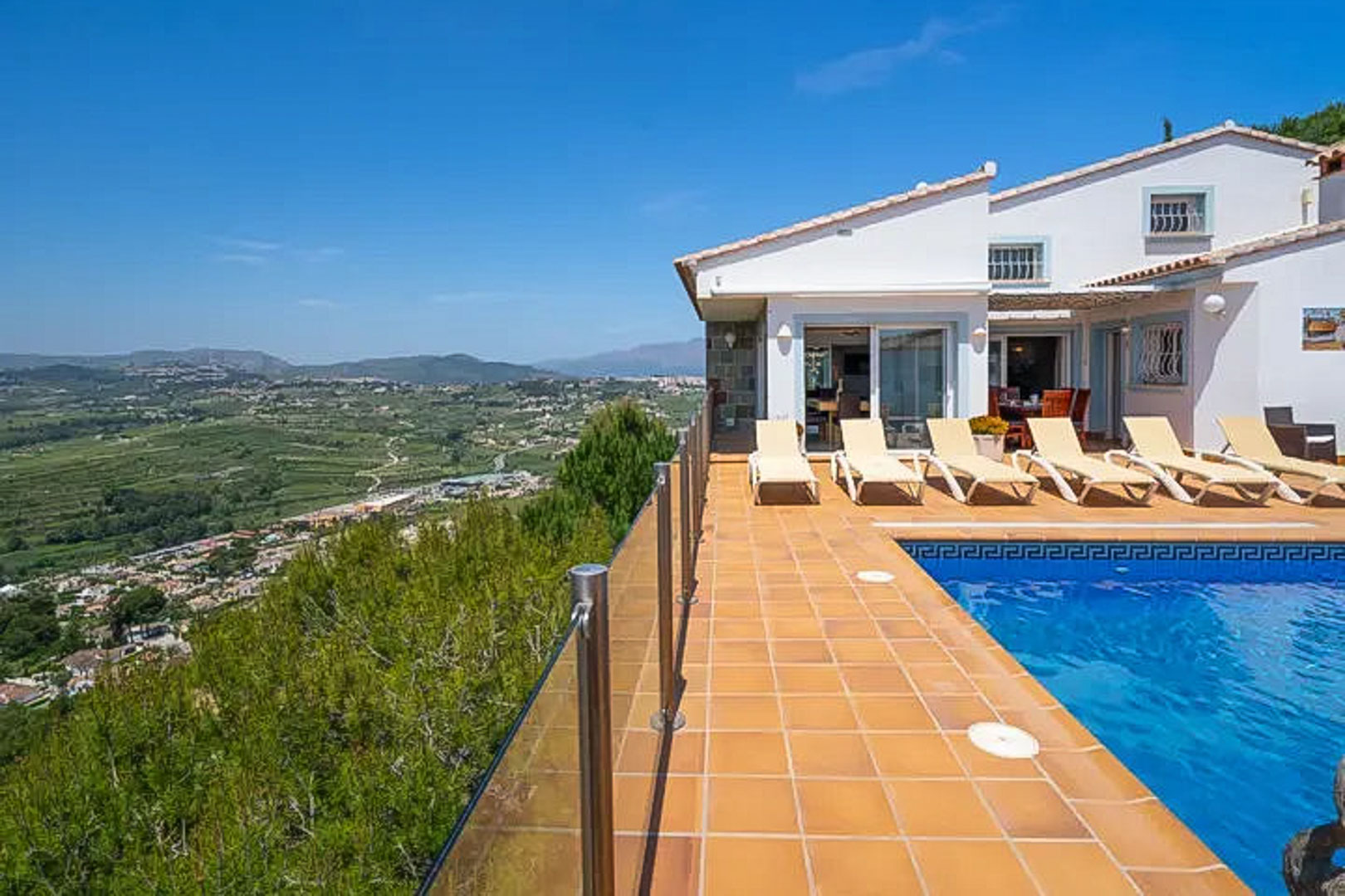 Erhöhte Terrasse mit blau gefliestem Pool, weißen Sonnenliegen und Glasgeländer mit weitem Blick über das Tal und die umliegenden Berge.