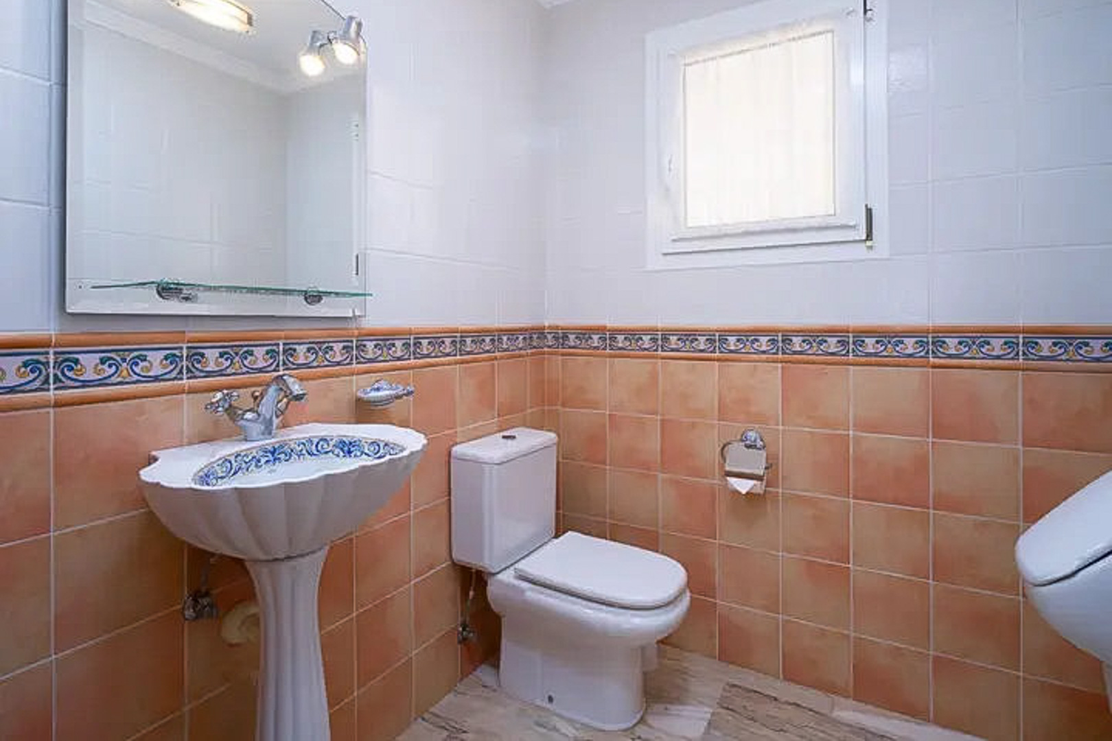 Gäste-WC mit dekoriertem Säulenwaschbecken, Wandfliesen in Terrakotta und Weiß mit Zierborte, großem Spiegel und kleinem Milchglasfenster.
