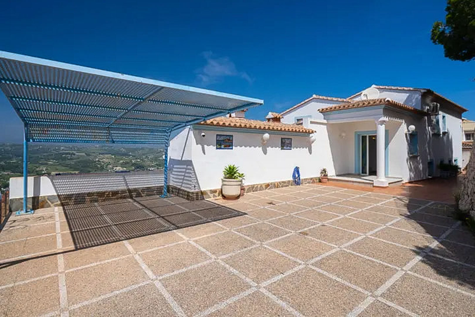 Großzügiger Außenparkplatz mit Fliesenboden, blauem Metall-Carport und weitem Blick über die hügelige mediterrane Landschaft und die Berge.