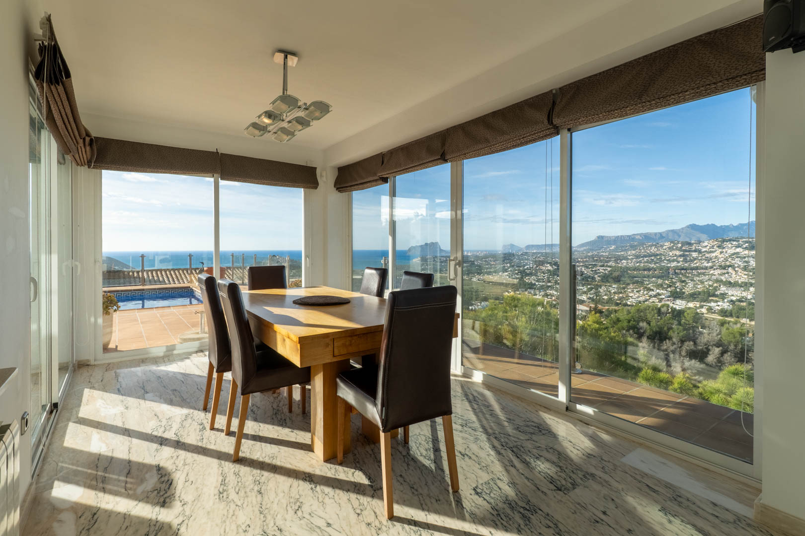 Heller Essbereich mit Marmorboden und massivem Holztisch. Bodentiefe Fenster bieten einen Panoramablick auf das Mittelmeer, den Peñón de Ifach und die umliegenden Berge.
