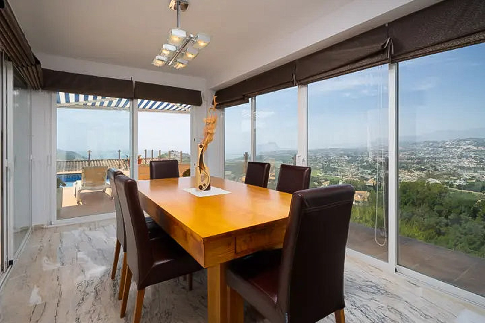 Heller Essbereich mit Massivholztisch und Ledersesseln, umgeben von großen Glasfronten mit Panoramablick auf das Meer und Zugang zur Poolterrasse.