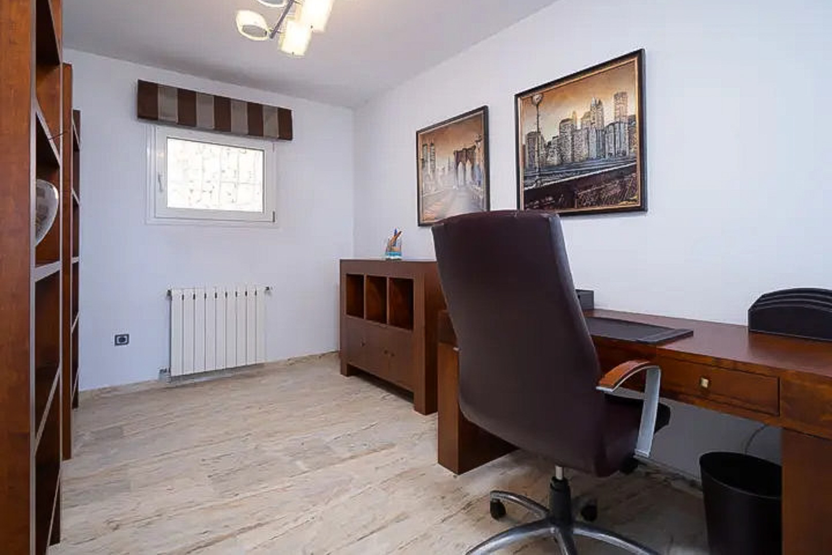 Helles Arbeitszimmer mit Holzmöbeln, funktionalem Schreibtisch und Ledersessel. Ausgestattet mit Regalen, hellem Natursteinboden und dekorativen Stadtbildern.