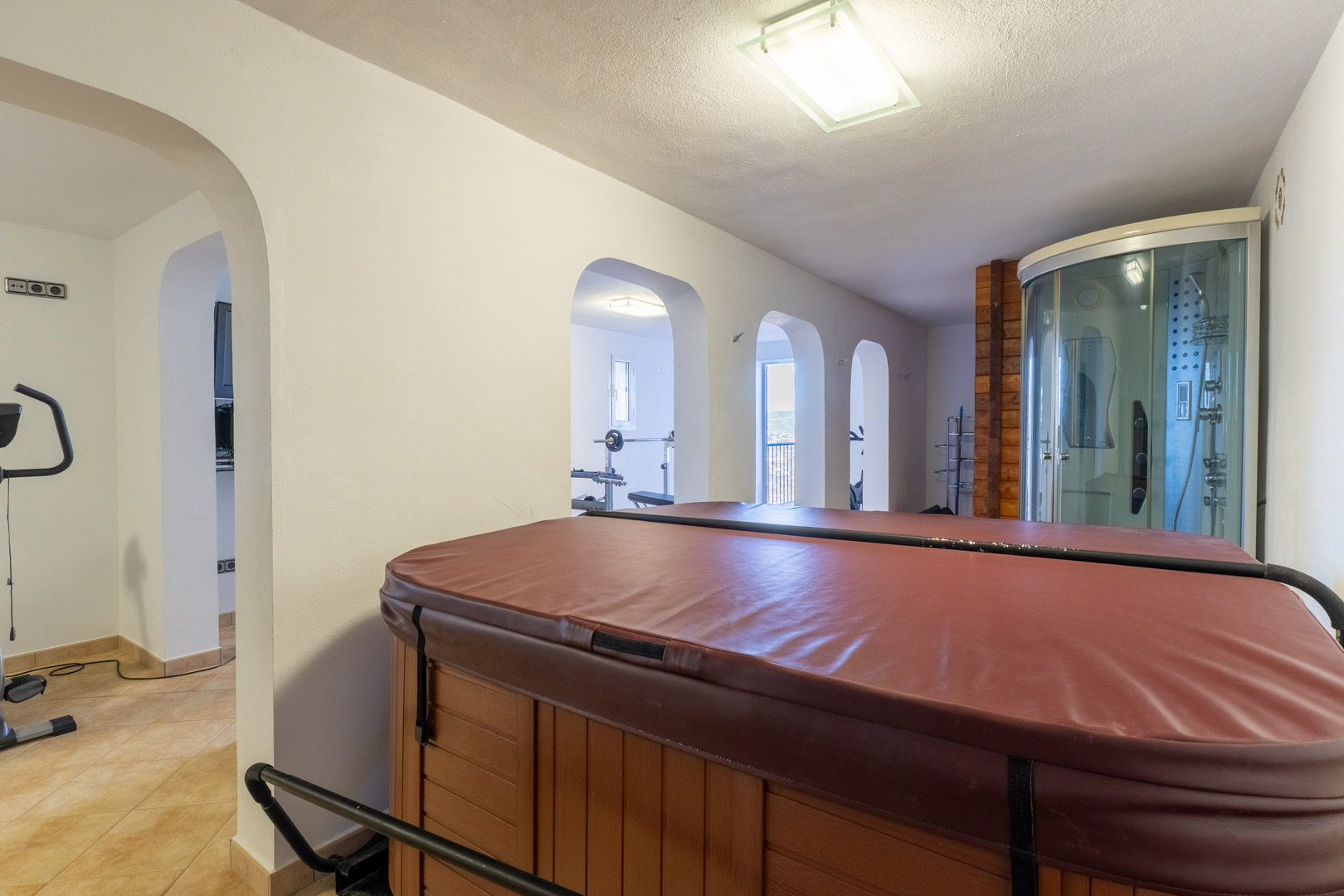 Indoor-Wellnessbereich mit abgedecktem Whirlpool, Hydromassage-Duschkabine und Rundbögen, die zum angrenzenden Fitnessraum führen.